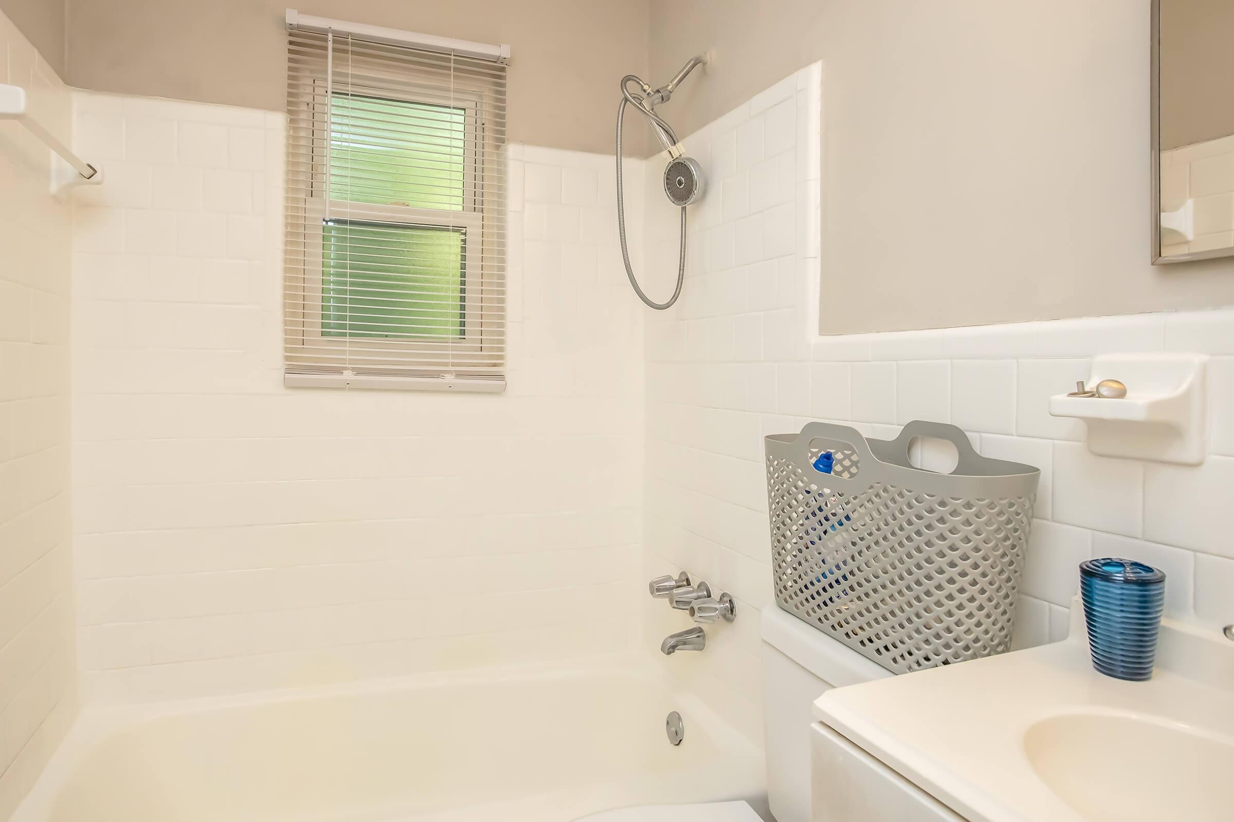 A clean and modern bathroom featuring a bathtub with a showerhead, white tiled walls, a window with blinds, and a countertop with a small blue glass. A gray storage basket is mounted on the wall next to the tub, making the space functional and organized.