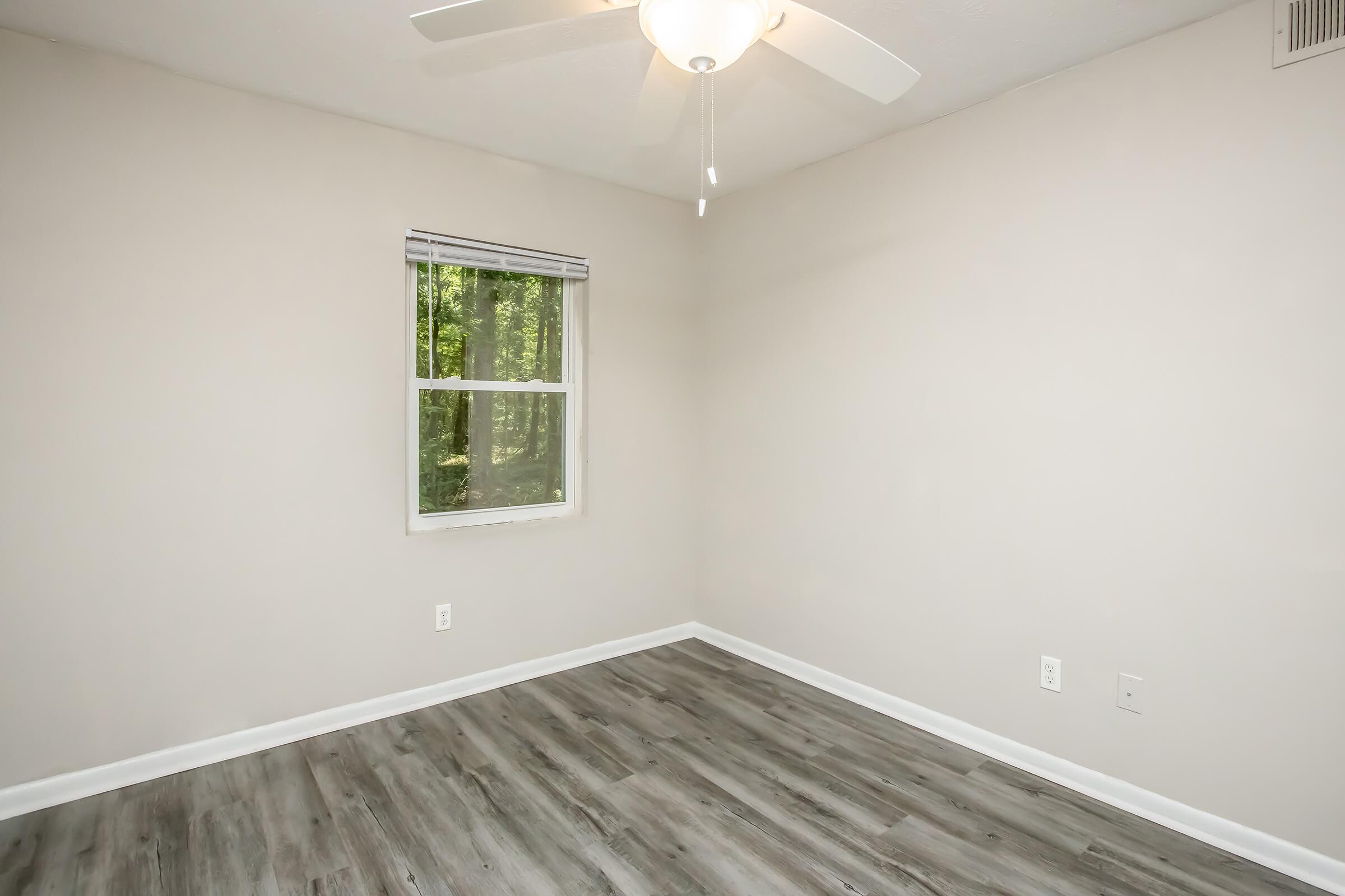 A minimalistic and empty room with light gray walls and a ceiling fan. There is a window revealing greenery outside, allowing natural light to enter. The floor is covered with light-colored laminate, adding to the room's airy atmosphere.