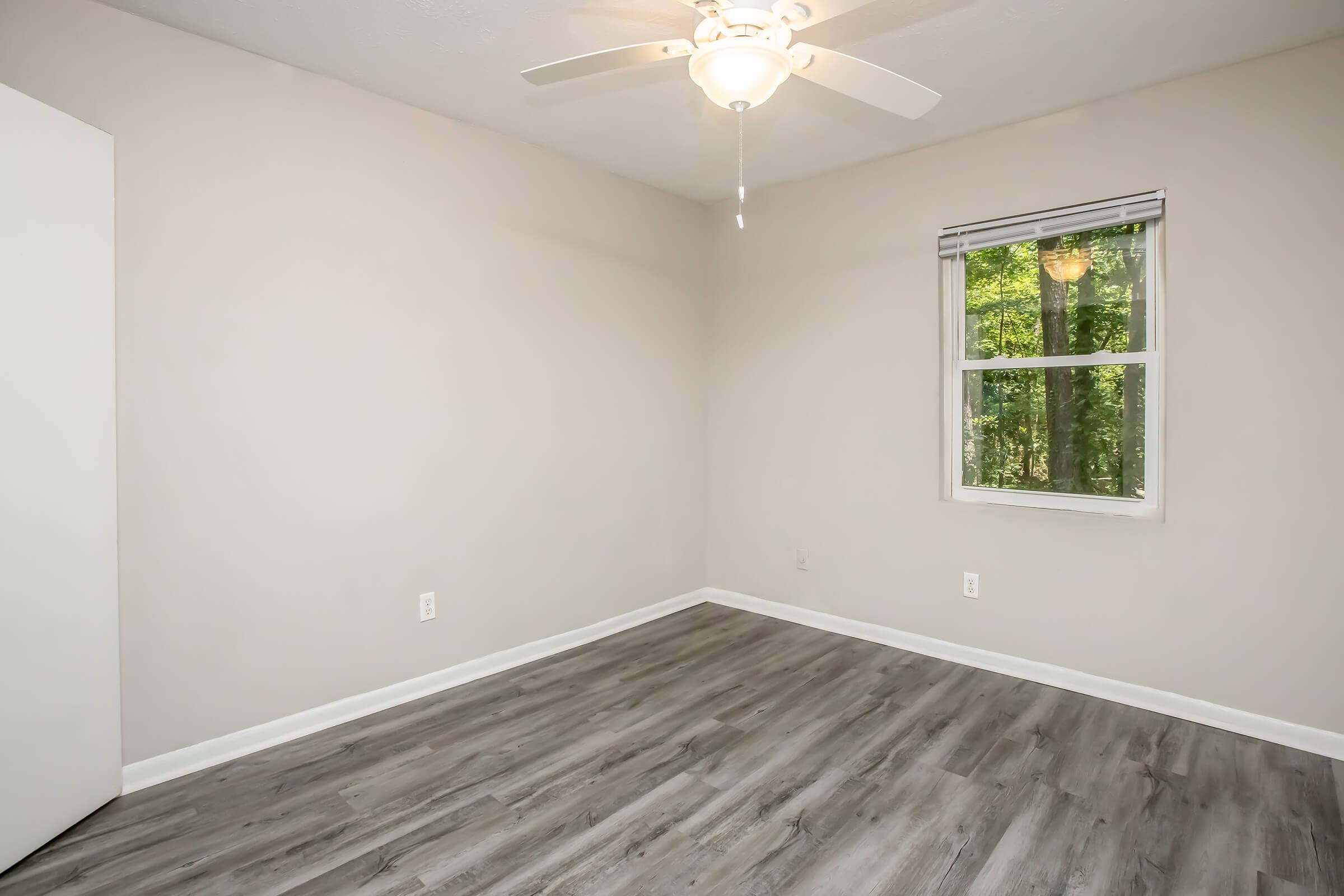 A well-lit, empty room featuring light gray walls and a ceiling fan. The floor has a wood-like finish, and there is a single window with a view of greenery outside. A closet door is visible on the left side of the image.