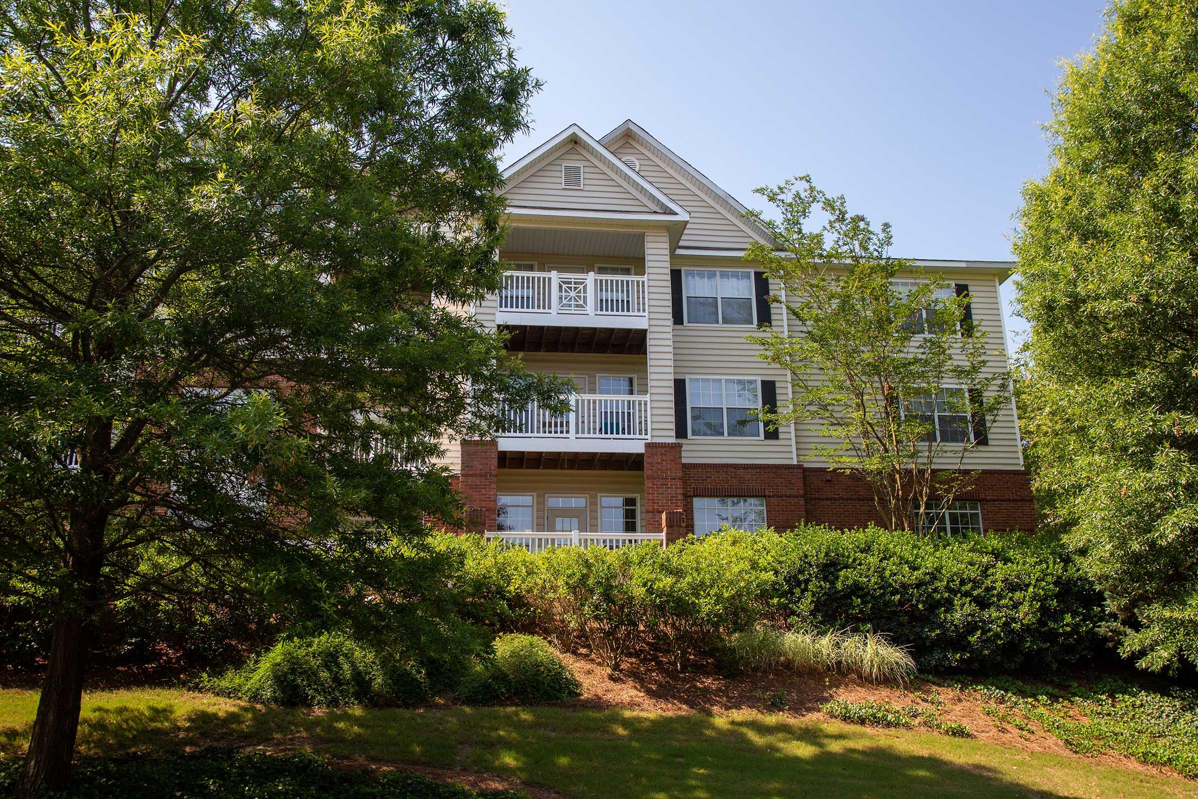 A multi-story residential building surrounded by greenery. The building features balconies with white railings and a mix of brick and siding exterior. Lush trees and shrubs are visible in the foreground, creating a serene environment. The sky is clear and blue, indicating a sunny day.