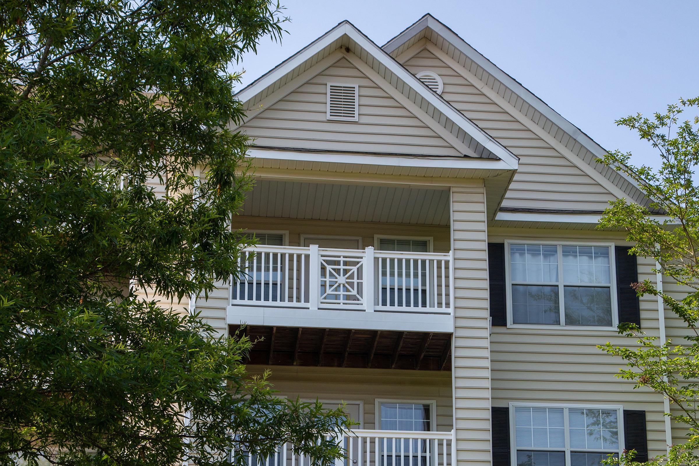 A close-up view of the upper floor of a beige apartment building featuring a balcony with white railings, surrounded by green trees. The building's exterior has shutters and windows that let in natural light on a clear day.