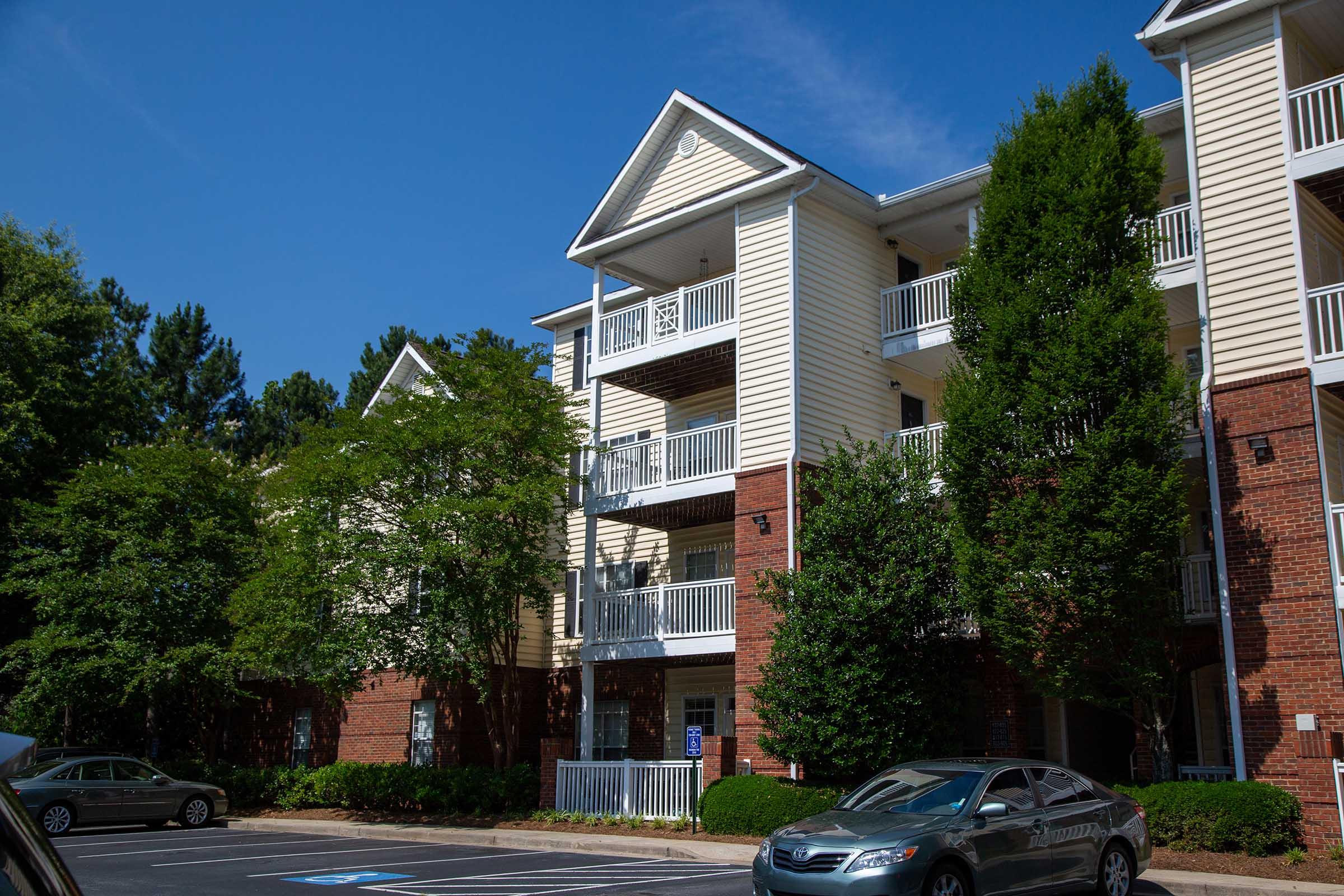 A multi-story apartment building featuring a mix of brick and light-colored siding. Balconies are visible on the upper floors, surrounded by lush greenery and trees. A parked car is in the foreground, with a clear blue sky above. The setting conveys a residential neighborhood atmosphere.