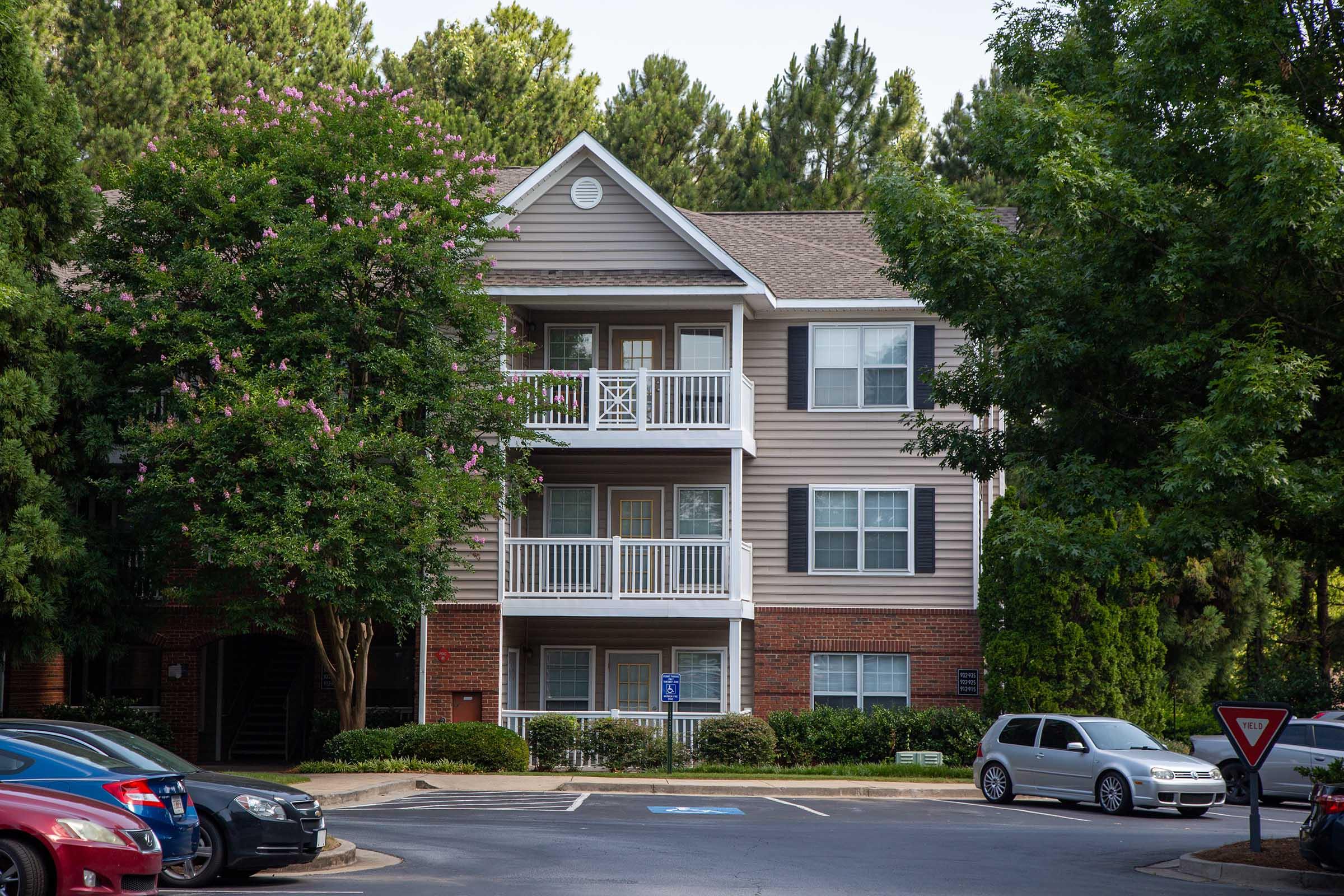 A three-story residential apartment building with a light brown exterior and white balconies. Surrounding the building are trees with green foliage and parked cars in a circular driveway. The setting appears to be in a suburban area with a clear sky.