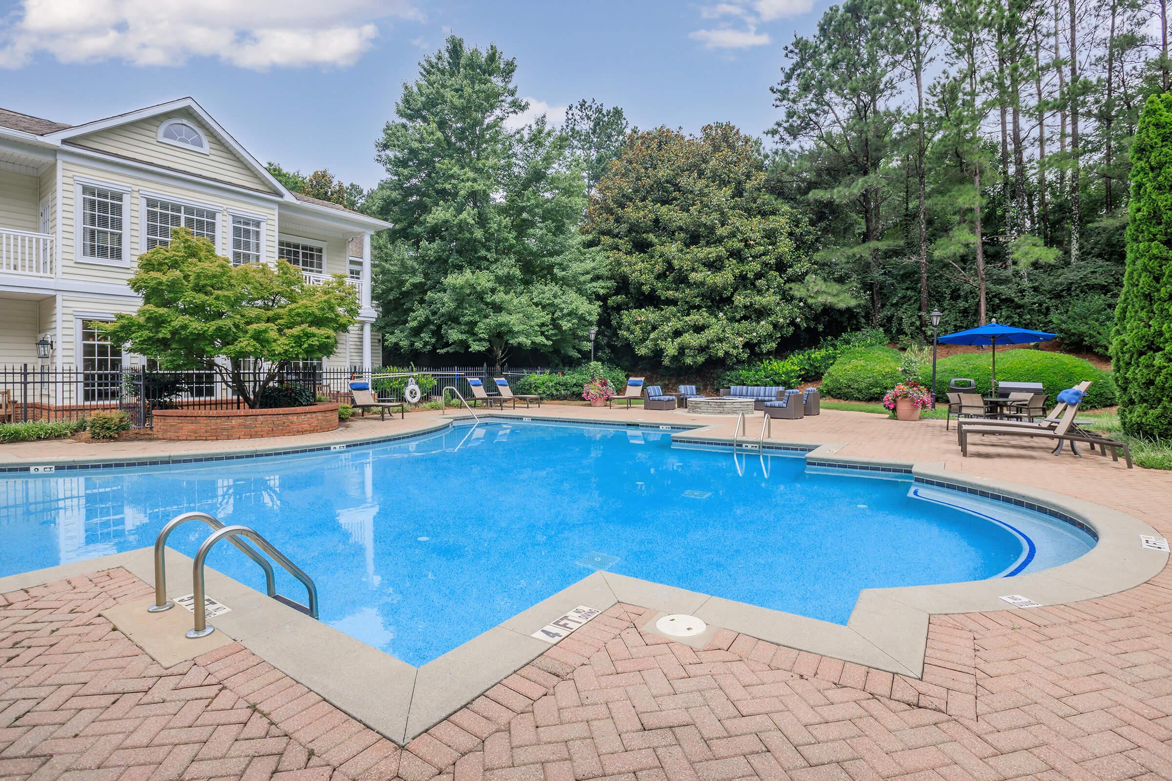 A serene outdoor pool area with clear blue water, surrounded by trees and greenery. Lounge chairs line the poolside, and a patio with umbrellas offers shaded seating. The brick-paved area enhances the inviting atmosphere, perfect for relaxation and leisure in a residential setting.