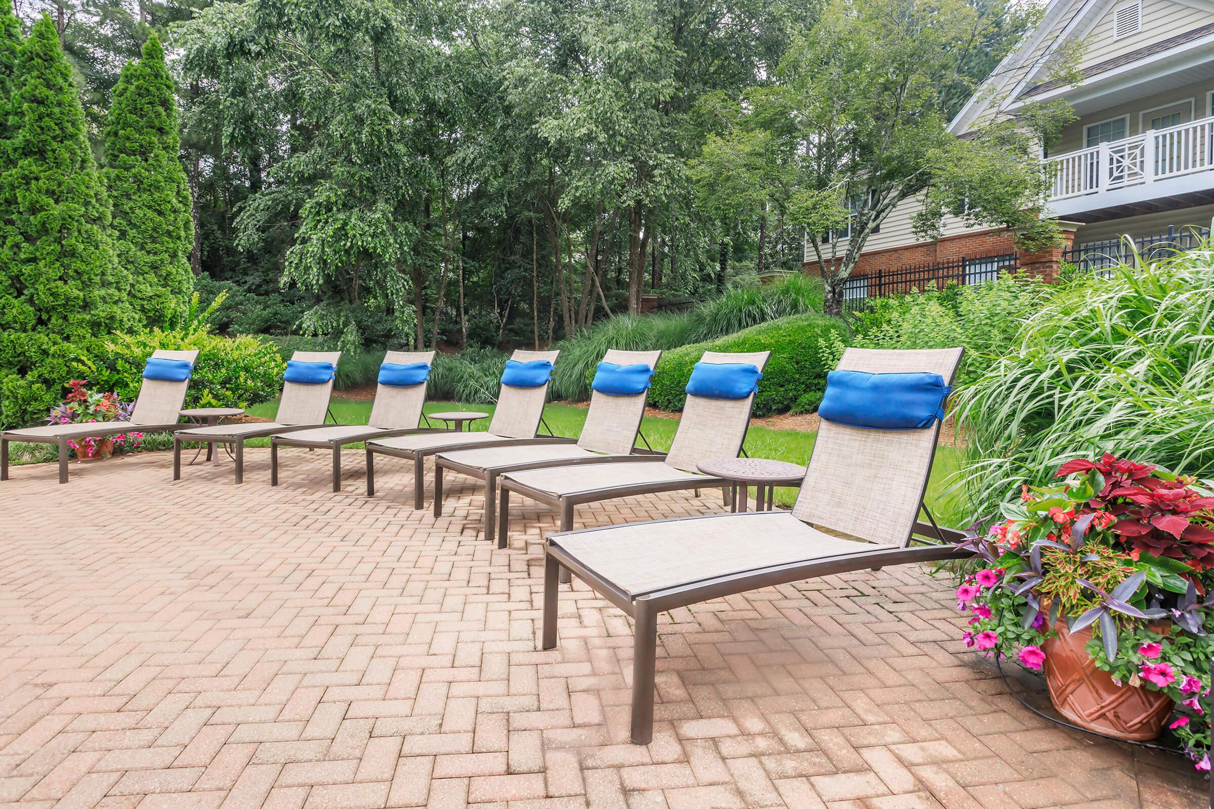 A serene outdoor space featuring a row of six lounge chairs with blue cushions on a brick patio, surrounded by lush greenery and colorful flowerbeds, creating a relaxing atmosphere near a residential building.