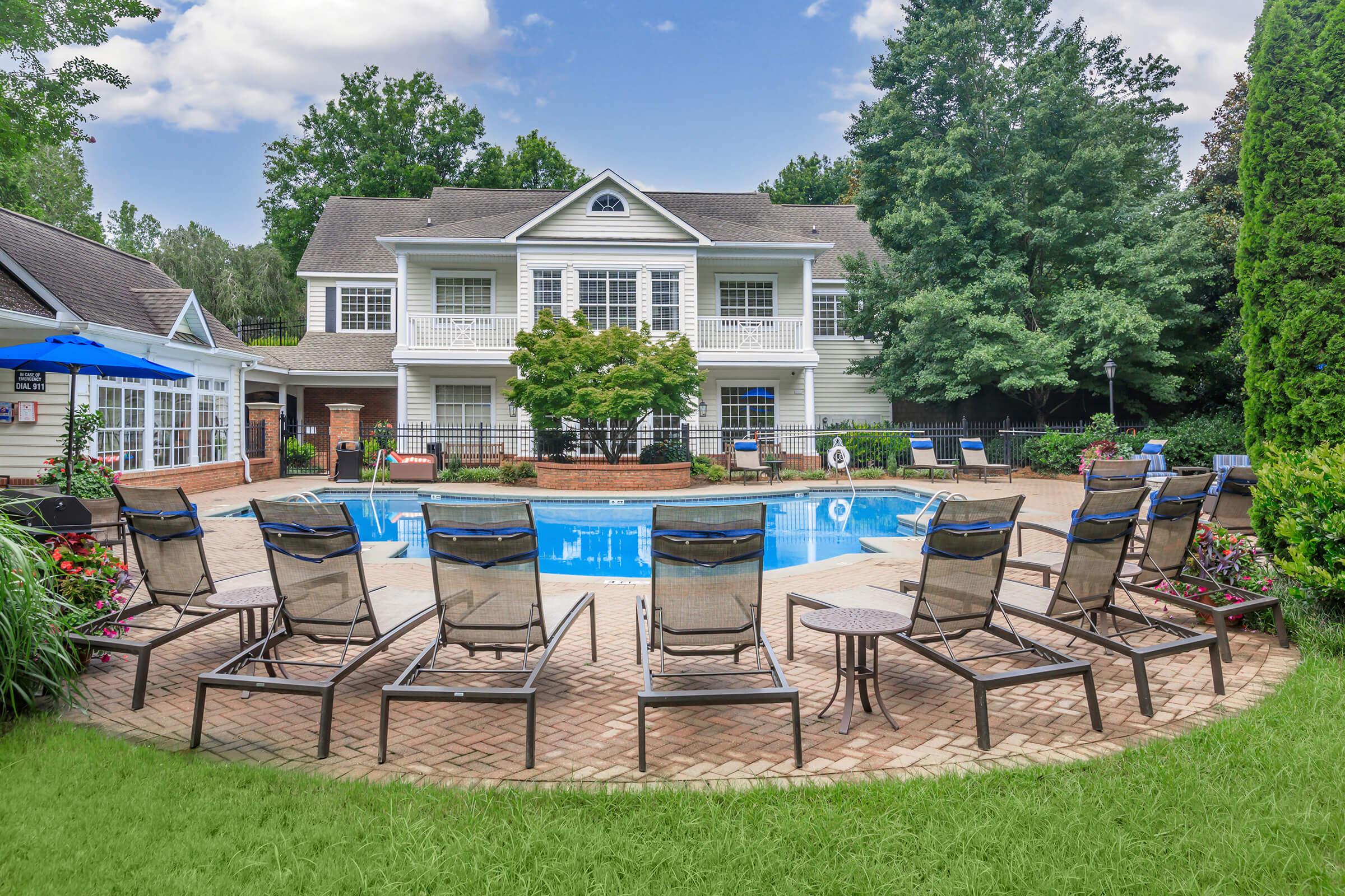 A tranquil outdoor pool area featuring a clear blue pool surrounded by lounge chairs on a paved deck. Lush greenery and flowering plants enhance the space, while a two-story building with multiple windows overlooks the pool. Sun umbrellas provide shade, creating a relaxing atmosphere for leisure and recreation.