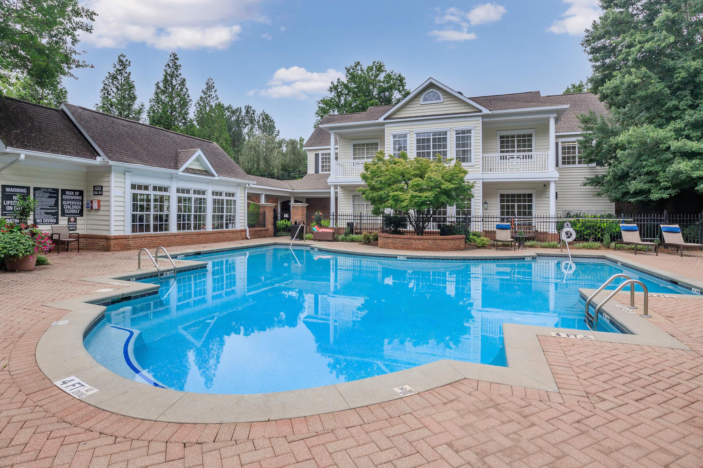 A clear blue swimming pool surrounded by a brick patio, with lounge chairs and lush greenery. In the background, there is a two-story building with a white facade and large windows, set against a partly cloudy sky. The area appears inviting and well-maintained, suitable for relaxation and leisure.
