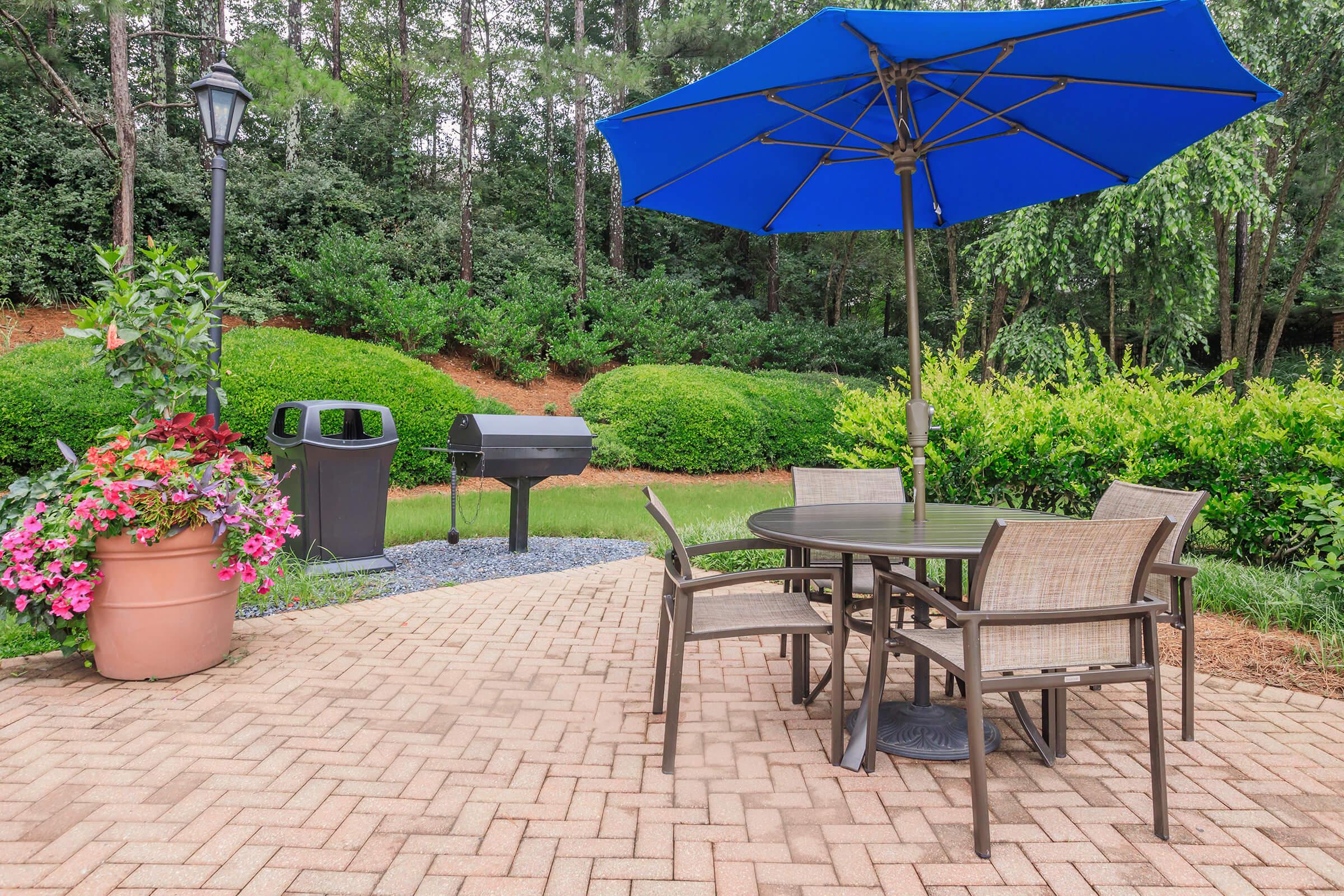A patio setting with a blue umbrella over a dining table surrounded by four chairs. In the background, there are neatly trimmed green bushes and a flowering pot. A barbecue grill is positioned nearby, and a lamp post adds to the outdoor ambiance. The paving stones create a neat, inviting area for gatherings.