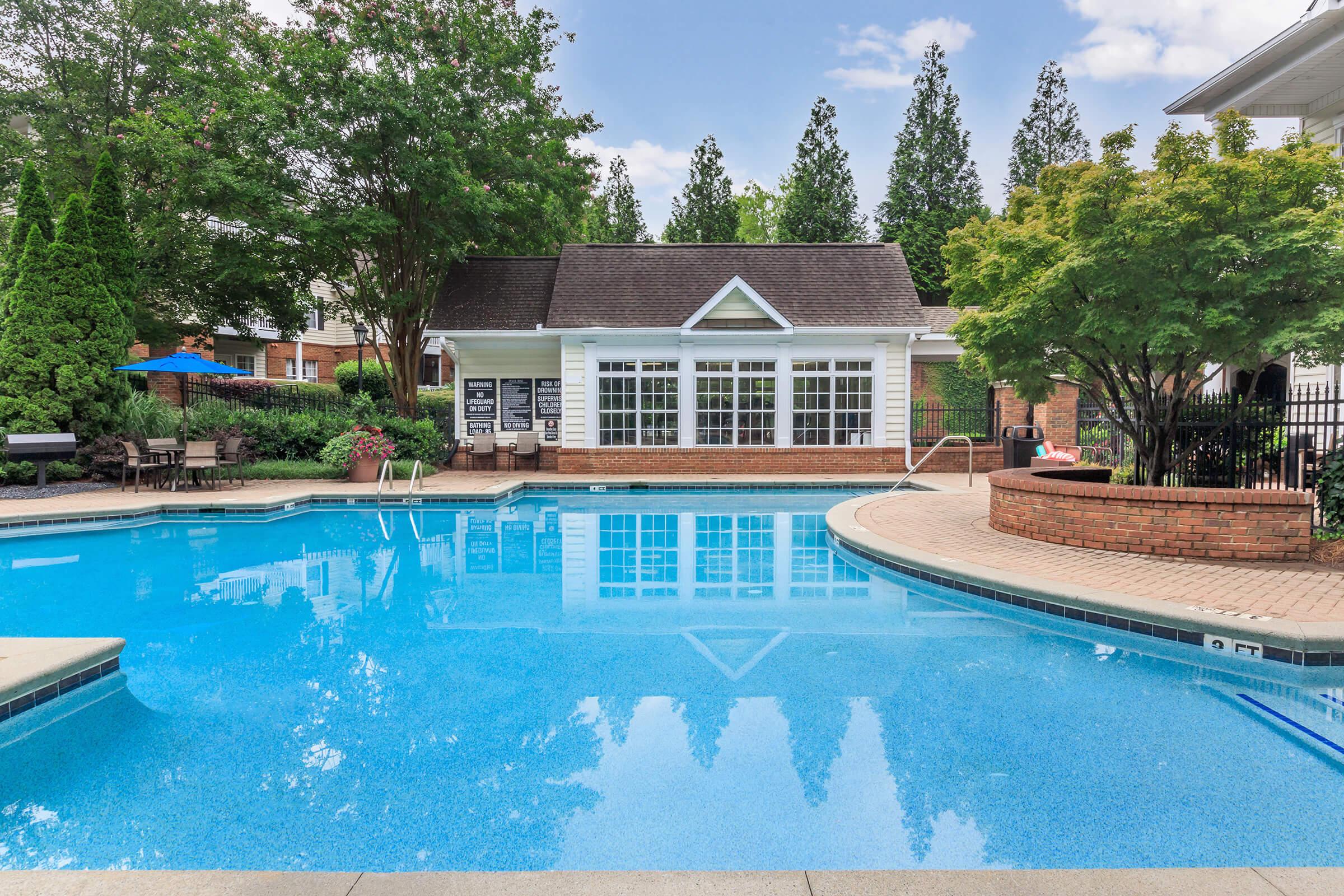 A clear blue swimming pool surrounded by a patio area, with lounge chairs and tables. In the background, there is a building with large windows and a sloped roof, along with trees and greenery providing a serene atmosphere. The sky is partly cloudy, adding a bright ambiance to the scene.