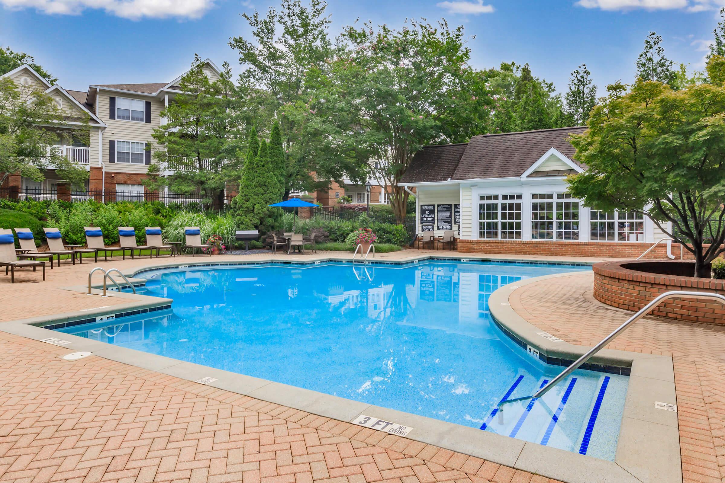 A clear blue swimming pool surrounded by lush greenery and residential buildings. There are lounge chairs arranged around the pool area, and a clubhouse with large windows is visible in the background. The setting is bright and inviting, ideal for relaxation and leisure activities.