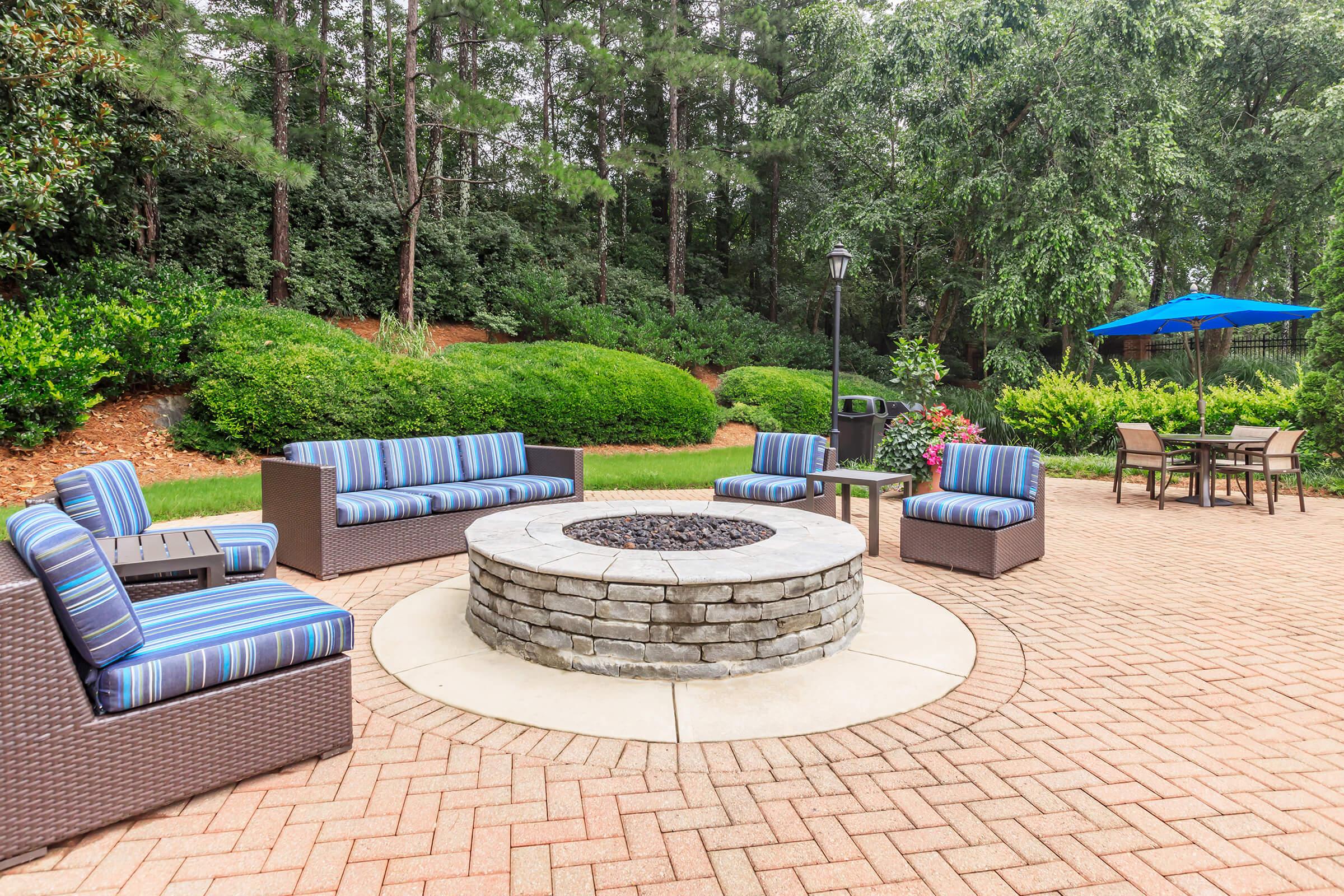 A cozy outdoor seating area featuring a circular stone fire pit surrounded by modern wicker furniture with striped cushions. Lush greenery and trees serve as a natural backdrop, complemented by a blue umbrella over a small dining table. The paved patio enhances the inviting atmosphere for relaxation and gatherings.
