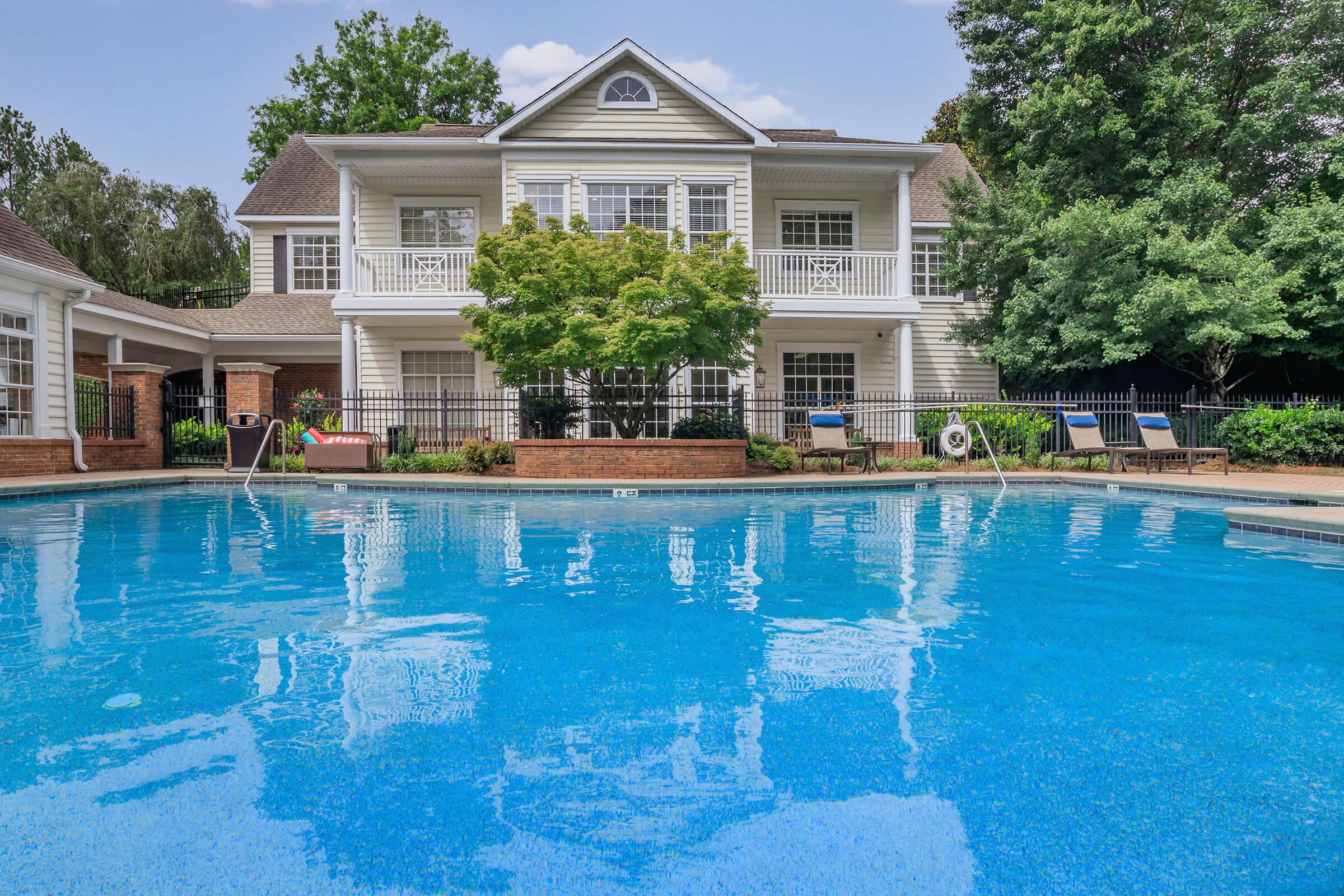A tranquil swimming pool with clear blue water in the foreground, surrounded by neatly trimmed greenery and lounge chairs. In the background, a spacious two-story building with a light-colored facade, balconies, and large windows, set against a clear sky and mature trees.