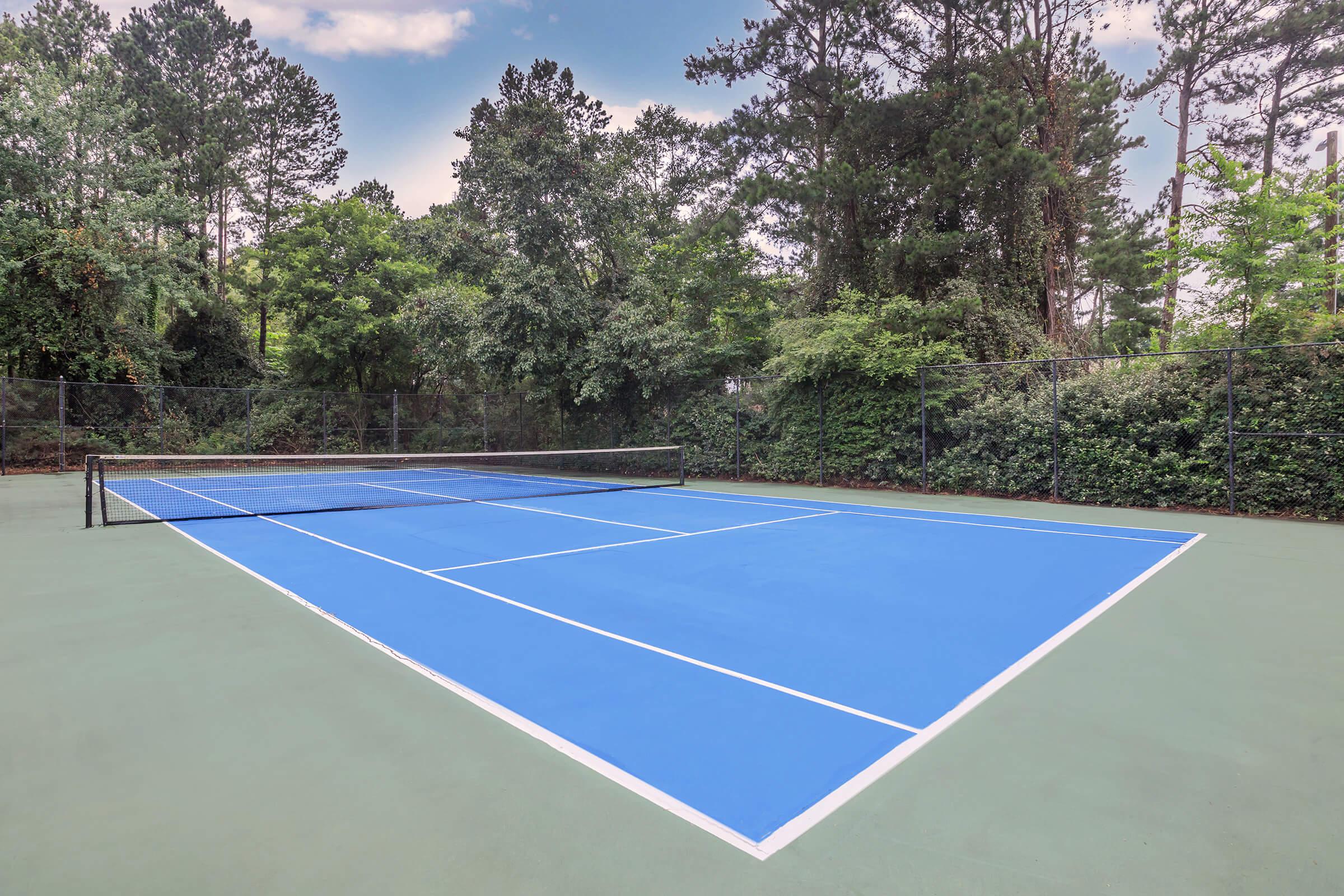 A vacant tennis court with a bright blue surface and white lines, surrounded by greenery and trees under a clear sky. The net is positioned at the center, and the court appears well-maintained, inviting for a game.