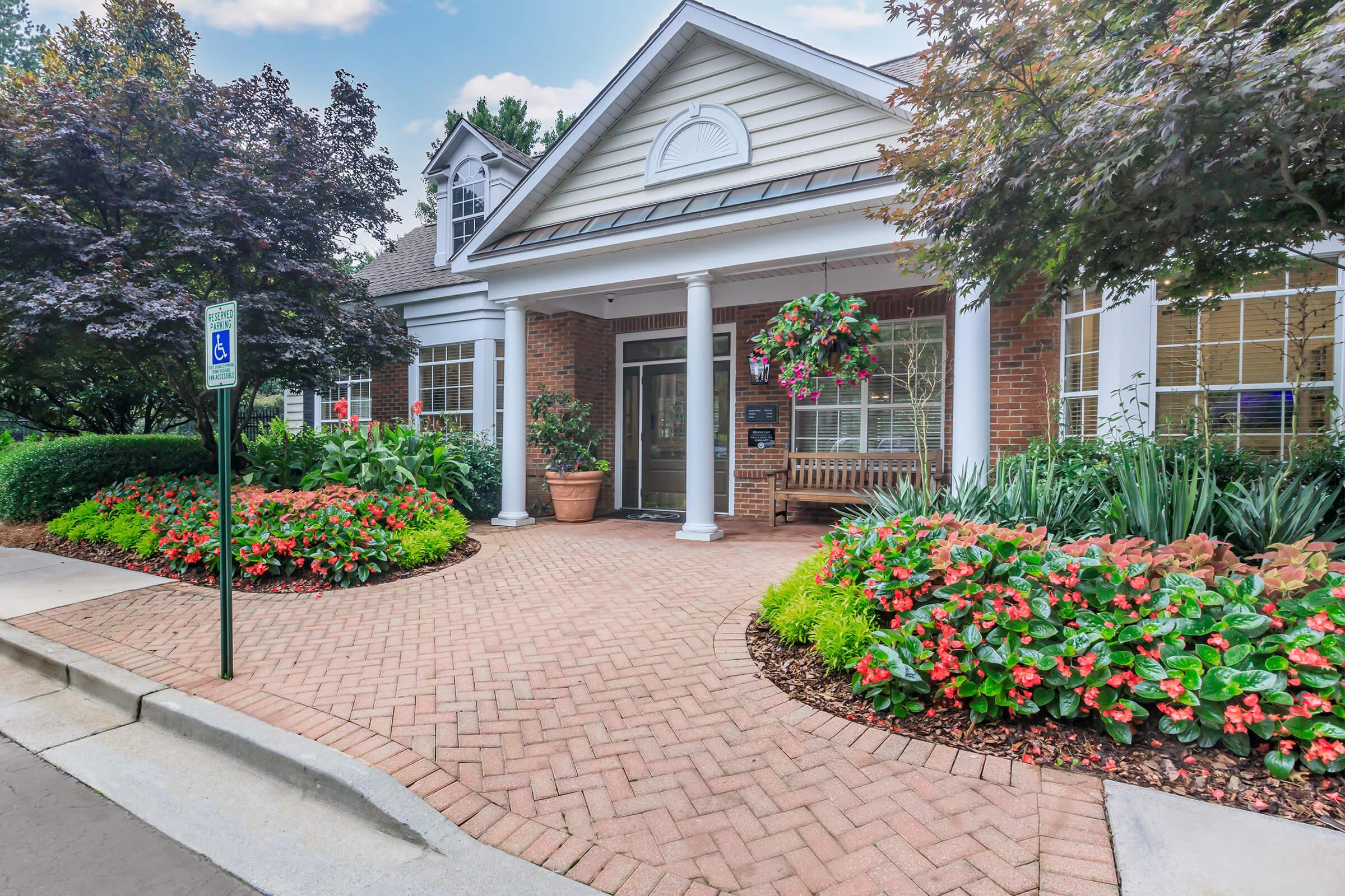 A stylish building entrance featuring a brick walkway leading to a covered porch. Colorful flower beds with red and pink blooms flank the pathway, while large potted plants and hanging baskets add greenery. The inviting atmosphere is enhanced by trees and shrubs surrounding the area.