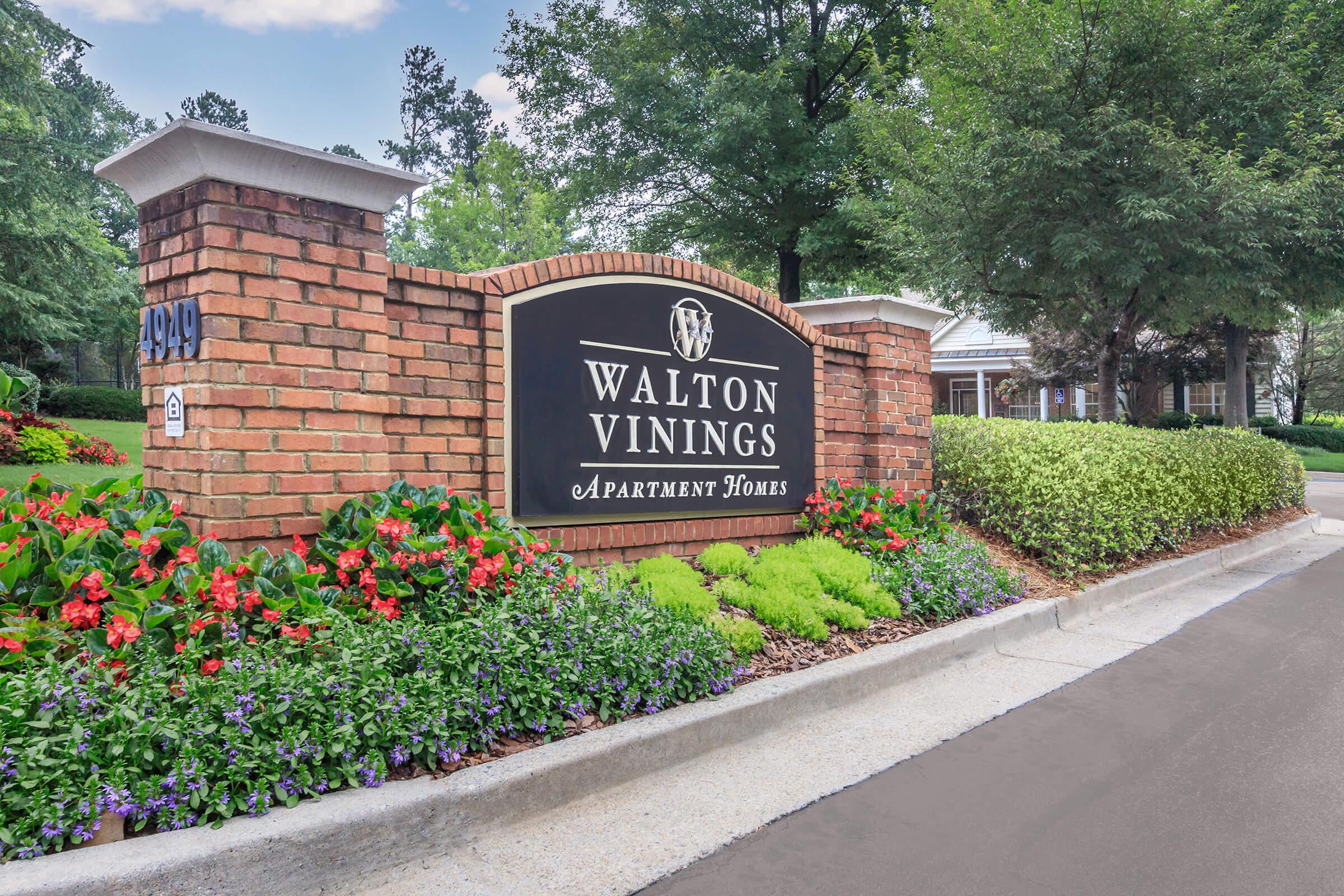 Sign displaying "Walton Vinings Apartment Homes" on a brick structure surrounded by colorful flower beds and greenery. The setting features a well-maintained entrance with trees in the background.