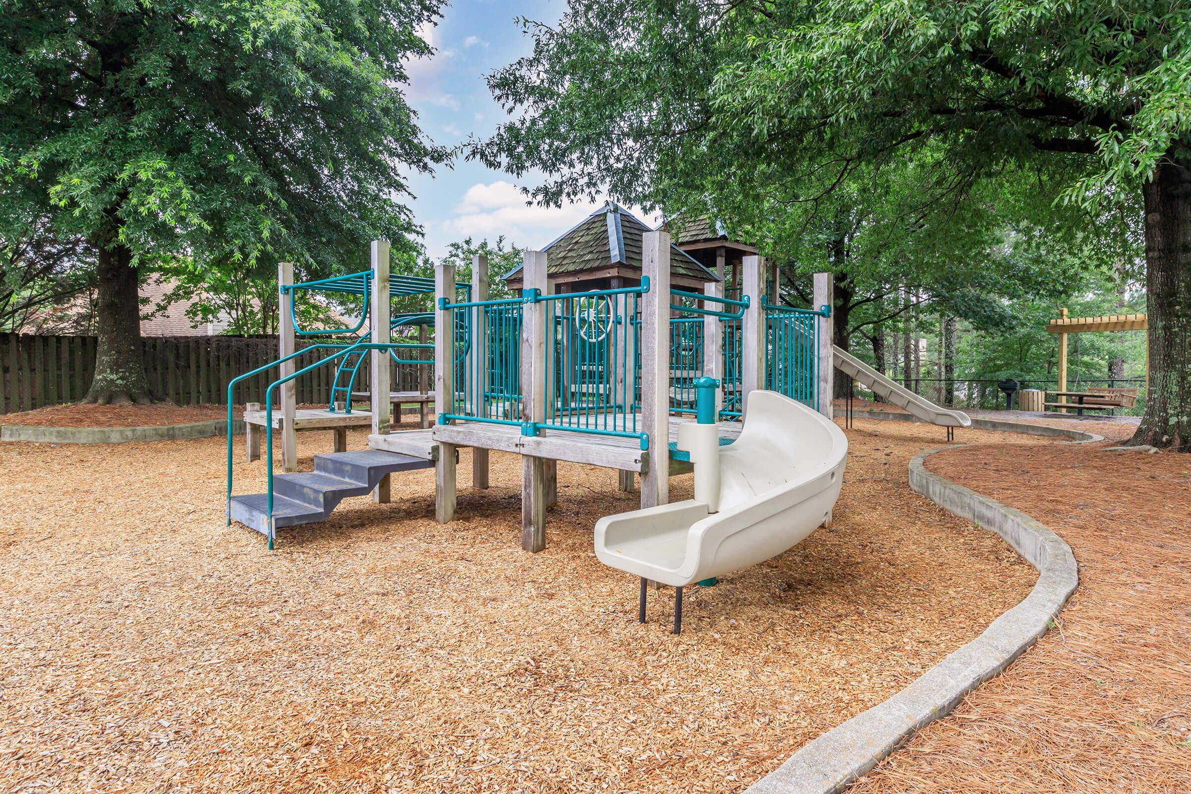 A playground featuring a wooden structure with slides, climbing equipment, and stairs, surrounded by trees and a bed of wood chips. The area is open and well-maintained, with picnic tables visible in the background, creating a welcoming outdoor space for children to play.