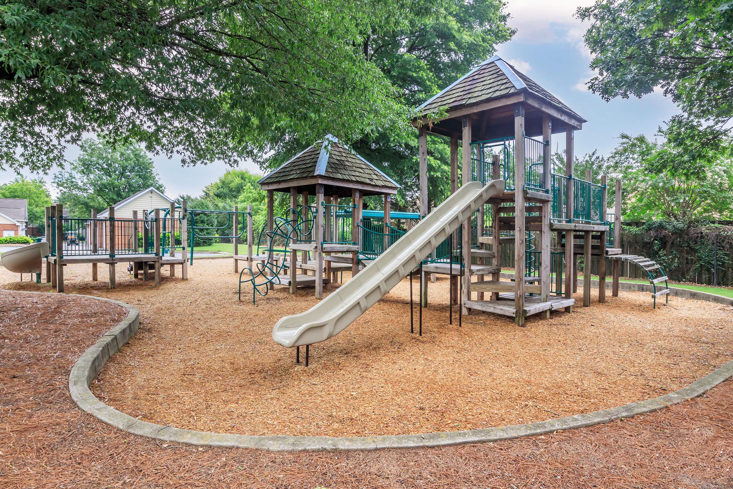 A wooden playground with two towers, a slide, and various climbing structures surrounded by soft mulch. Lush green trees provide shade, and a nearby fence separates the area from a residential neighborhood.