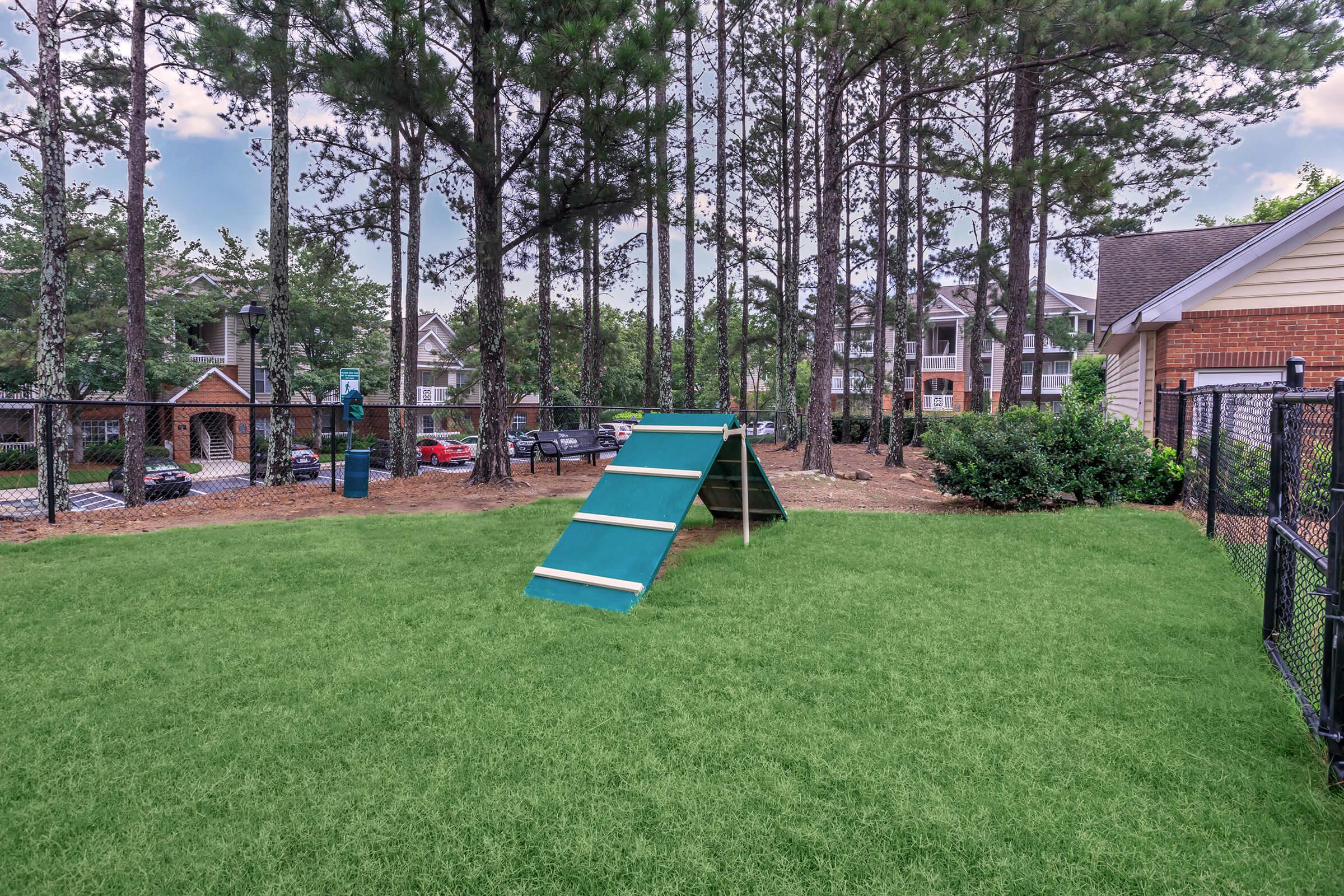 A grassy area with an artificial turf surface featuring a teal climbing structure, surrounded by tall pine trees. In the background, there are apartment buildings and a fenced area. The scene is bright and inviting, ideal for outdoor activities or play.