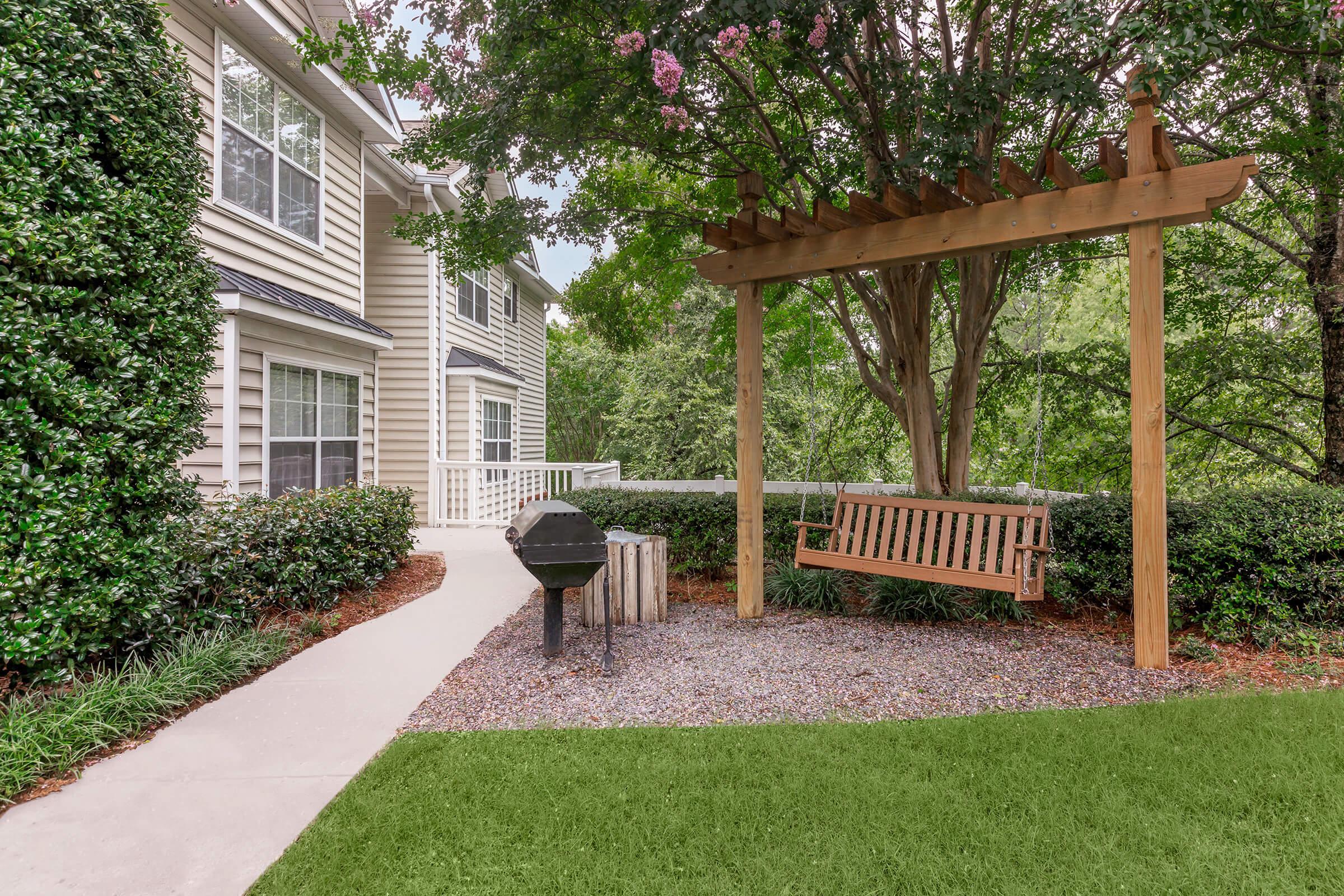A peaceful outdoor area featuring a wooden swing beneath a pergola, a charcoal grill nearby, and landscaped greenery, including low bushes and trees. The setting is adjacent to a residential building with a light-colored facade and large windows.