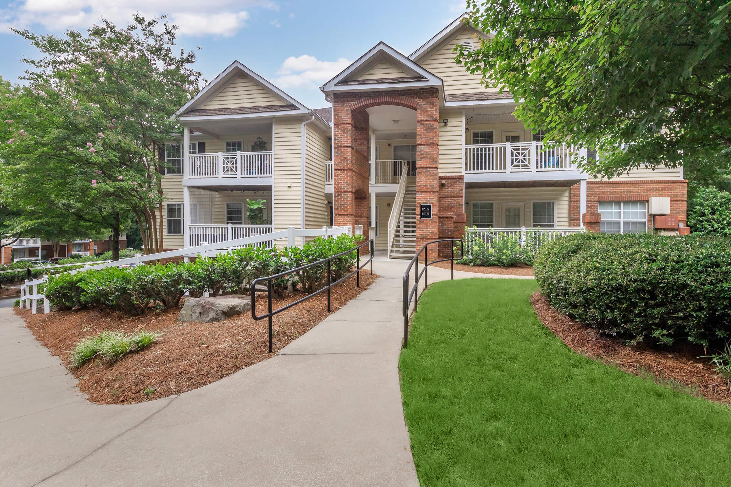 Two-story residential building with a mix of light-colored siding and brick accents. The entrance features a covered porch with steps leading up to it. Surrounding the building are landscaped areas with green grass, shrubs, and trees, creating a welcoming environment. Walkways connect the entrance to the surrounding pathways.