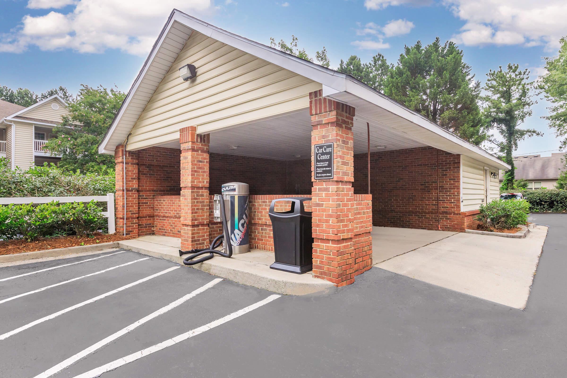 A covered car wash area with a vending machine and vacuum station, surrounded by trees and shrubs. The structure features brick walls and white siding, with a clear sky above. Parking spaces are marked in front, indicating designated spots for car washing.