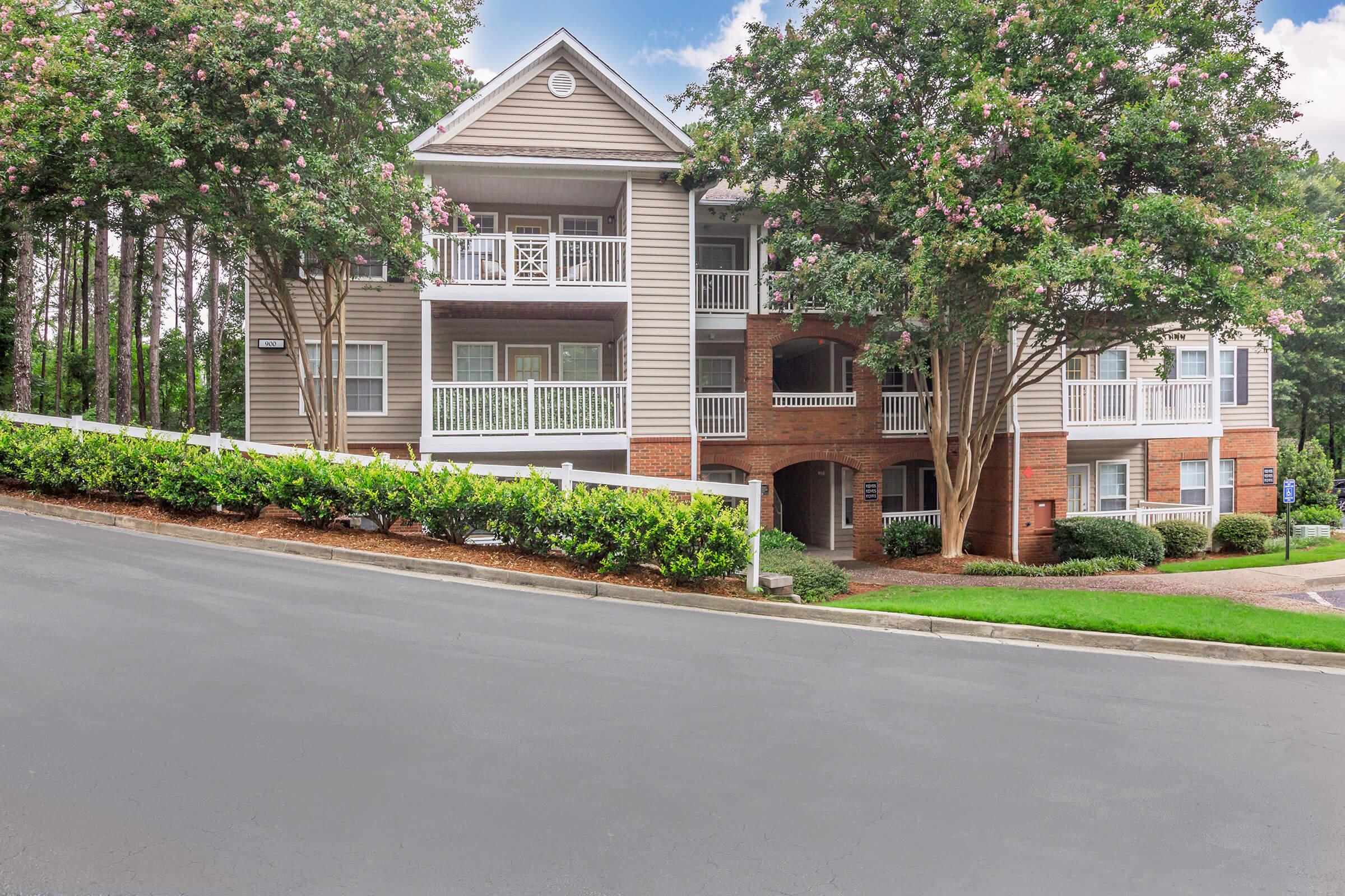 A three-story apartment building with balconies, surrounded by trees and flowering shrubs. The building features beige siding and red brick accents, set on a sloped driveway leading to a landscaped area with green bushes. The sky is partly cloudy, adding a serene atmosphere to the scene.
