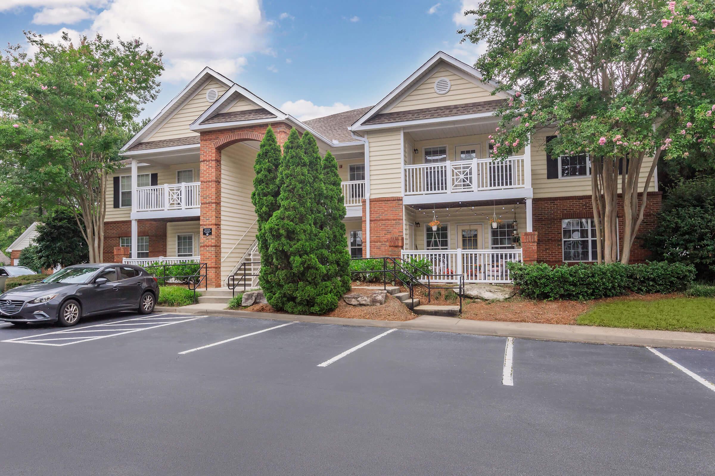 A two-story apartment building featuring a mix of brick and siding exterior, surrounded by trees and shrubs. A black car is parked in the foreground on a paved lot, with several parking spaces visible. The sky is partly cloudy, creating a pleasant and inviting atmosphere.