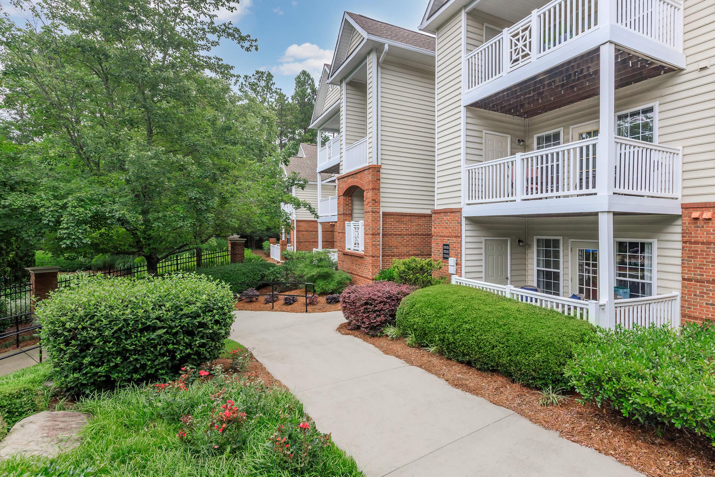 A walkway leading to a multi-story residential building with balconies. The surroundings feature well-maintained greenery, including shrubs and small flowering plants, against a clear blue sky. The architecture includes brick and light-colored siding, creating a welcoming atmosphere.