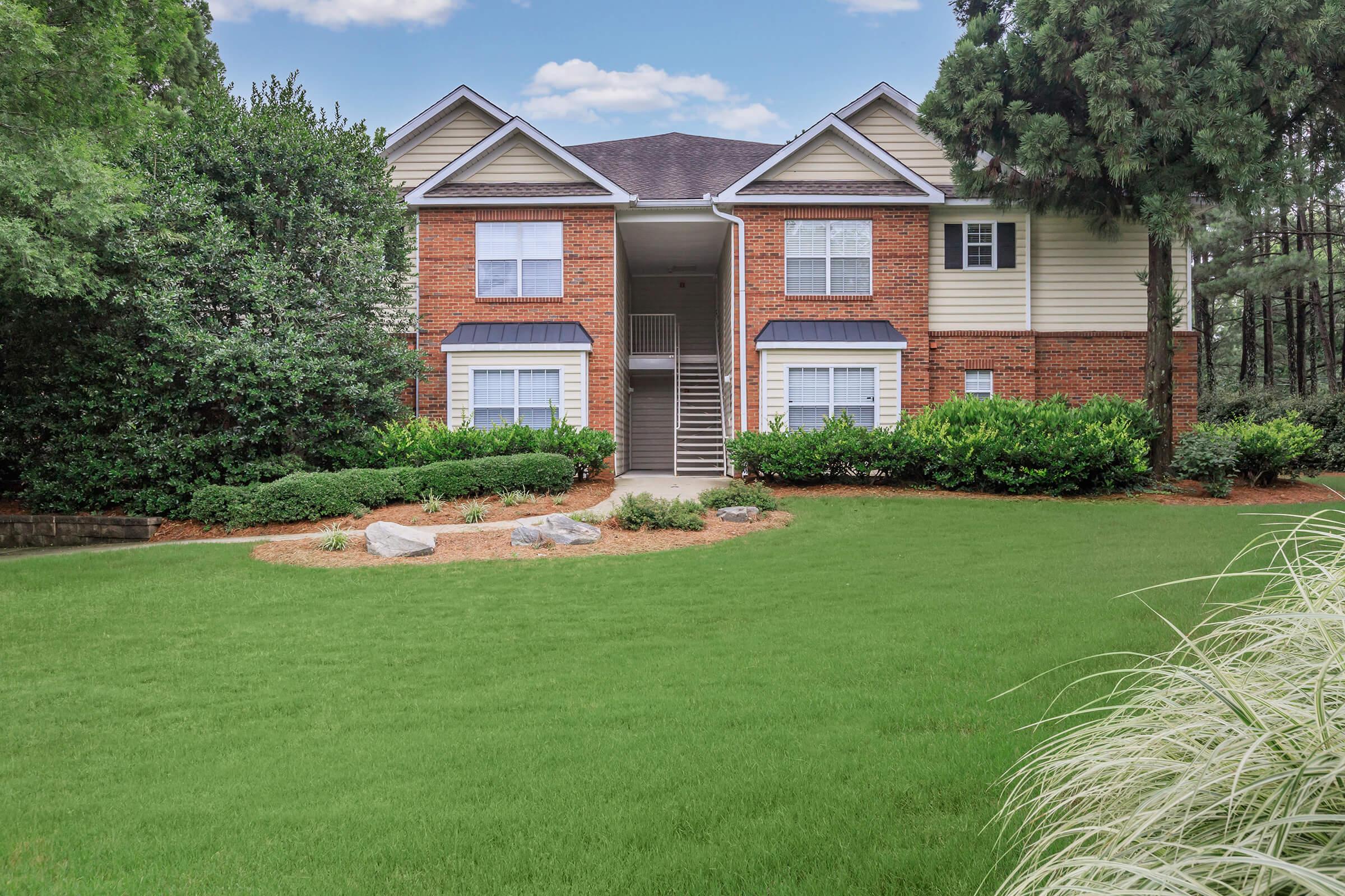 An exterior view of a two-story residential building, featuring a brick facade and light-colored siding. The building is surrounded by well-maintained green lawns, shrubs, and trees. A pathway leads to the entrance, and the sky is partly cloudy.