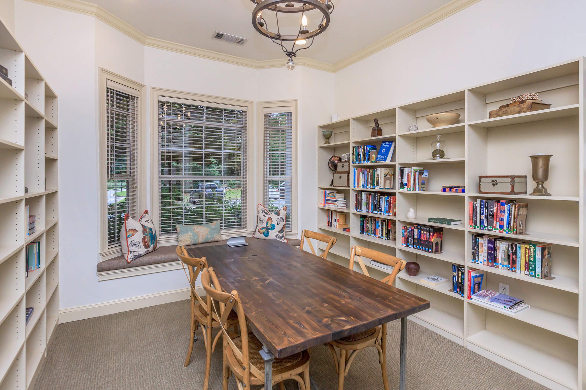 A cozy room featuring a wooden table surrounded by chairs, with bookshelves filled with books on either side. Large windows allow natural light, and decorative pillows are visible on a window seat, creating a warm and inviting atmosphere for reading or meetings.