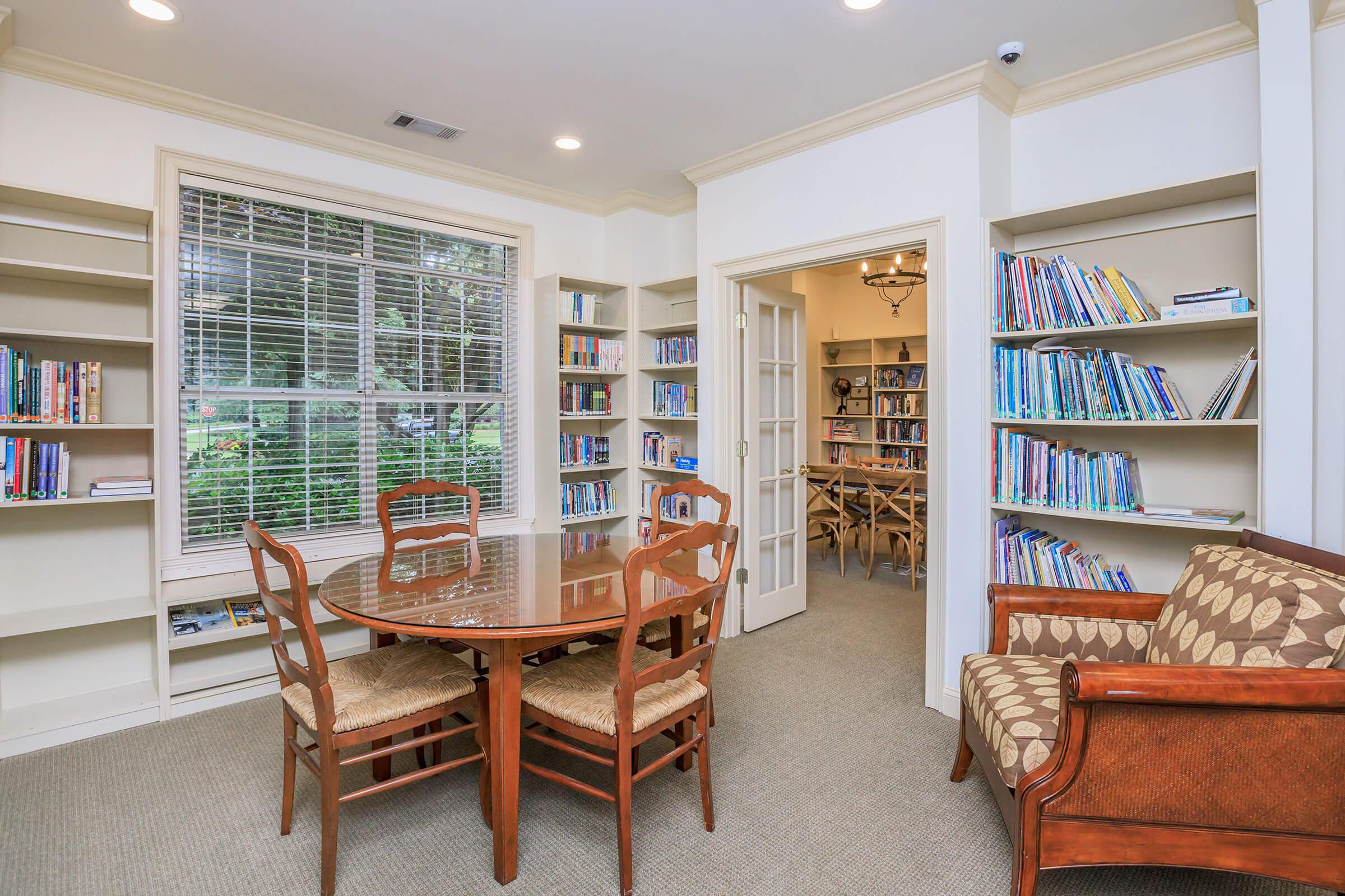 A cozy library space featuring a round wooden table with four chairs, surrounded by bookshelves filled with various books. A comfortable sofa with a patterned fabric sits against the wall. Natural light flows through a large window, illuminating the room and creating a welcoming atmosphere.