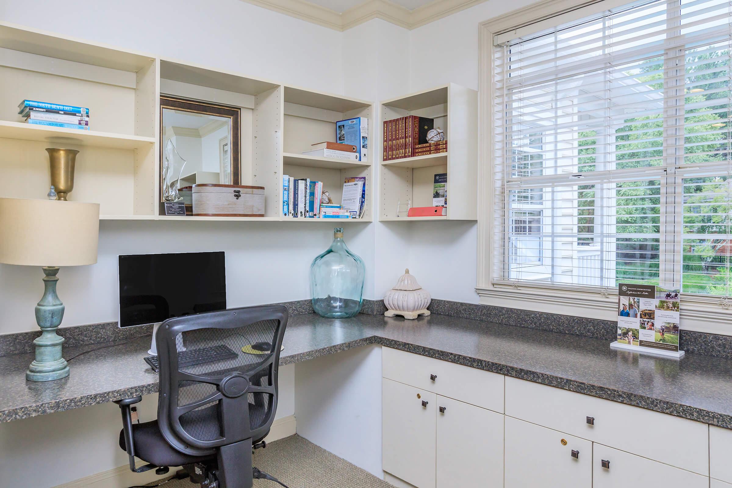 A well-organized home office featuring a corner desk with a computer monitor, a lamp, and decorative items. Shelves above hold books and personal items, while a window allows natural light to fill the space, creating a bright and inviting atmosphere.