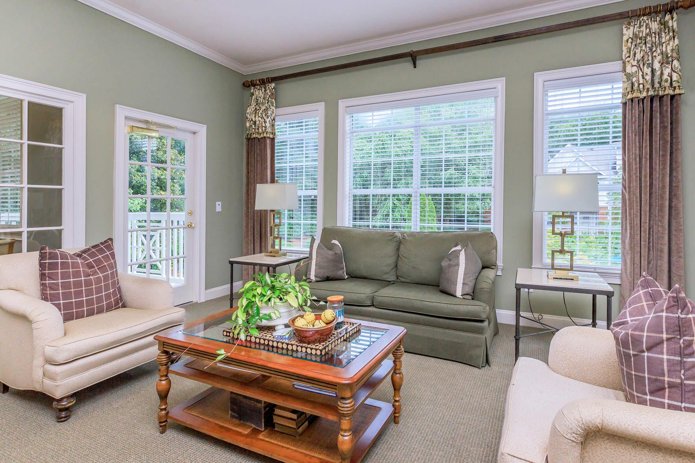 A cozy living room featuring a green wall, large windows with curtains, a coffee table with decorative items, and comfortable seating, including a sofa and armchairs. The space is well-lit and inviting, with a view of greenery outside.