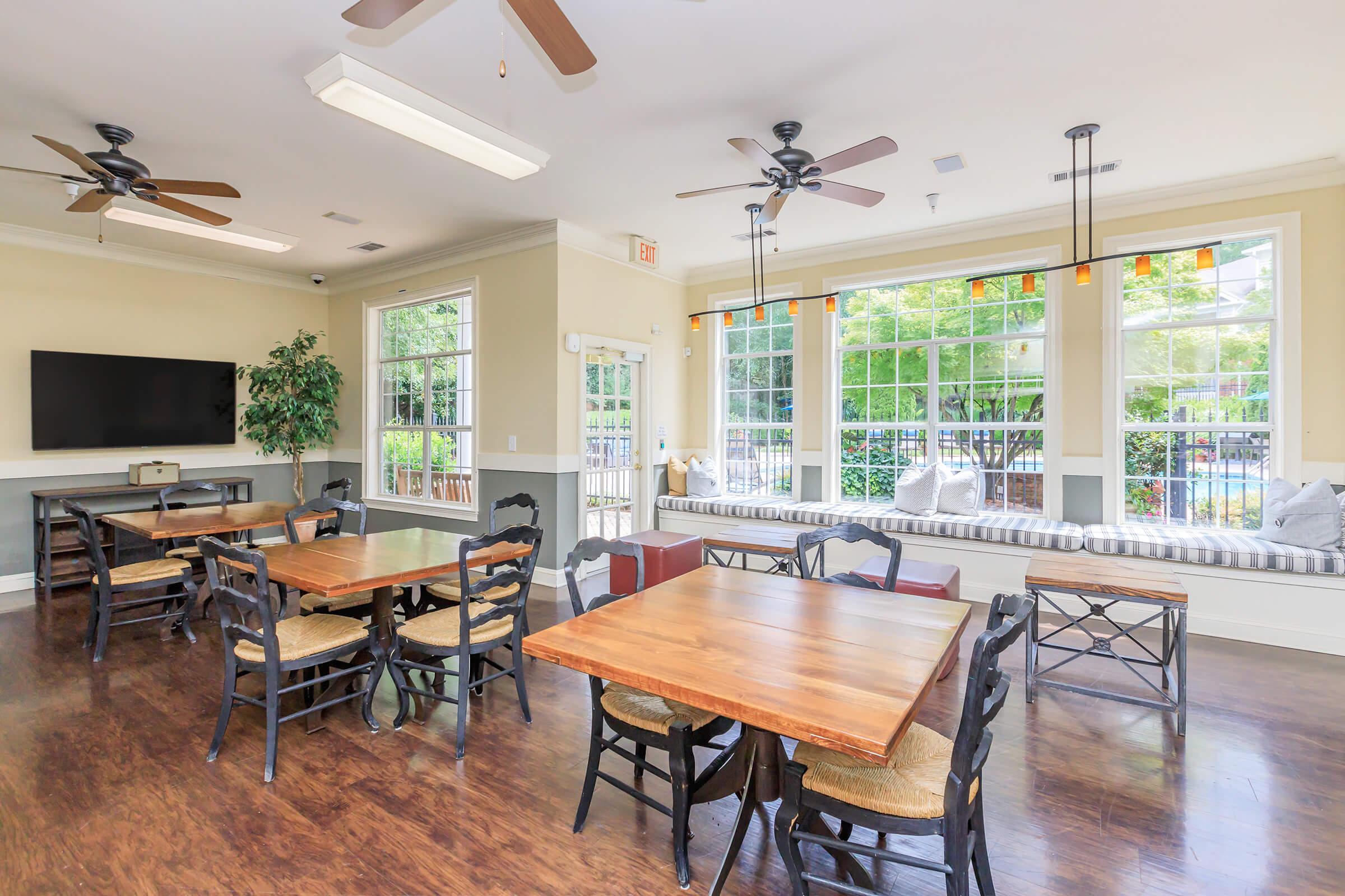 A well-lit common room featuring six wooden dining tables with chairs, ceiling fans, large windows with greenery outside, and a cozy window nook with seating and decorative pillows. A television is mounted on the wall, enhancing the inviting atmosphere of the space.