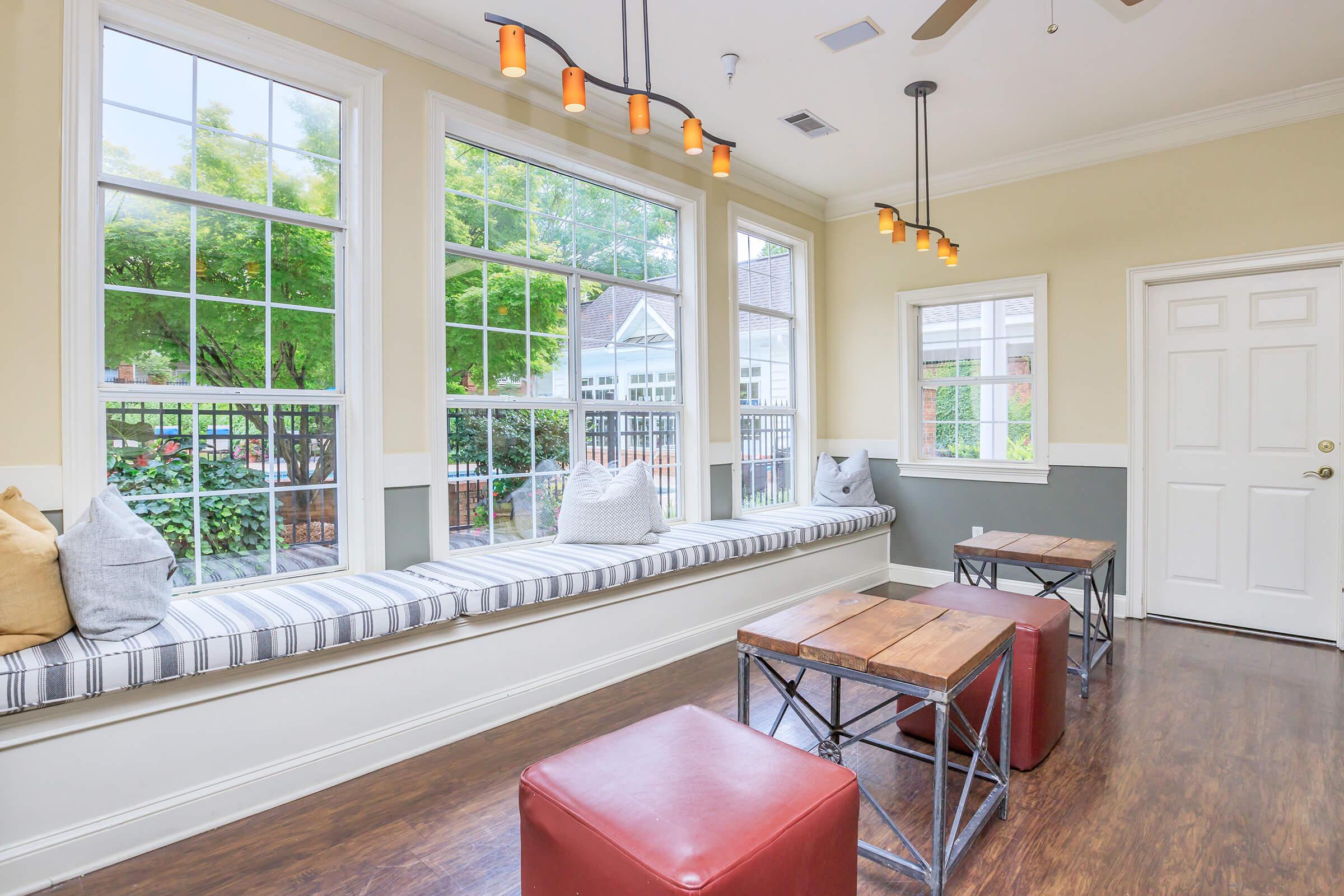 A bright and inviting room featuring large windows with greenery outside. There is a cozy window seat with striped cushions, complemented by wooden tables. The space has a warm atmosphere, enhanced by pendant lighting and a contemporary color scheme of soft neutrals and dark wood flooring.