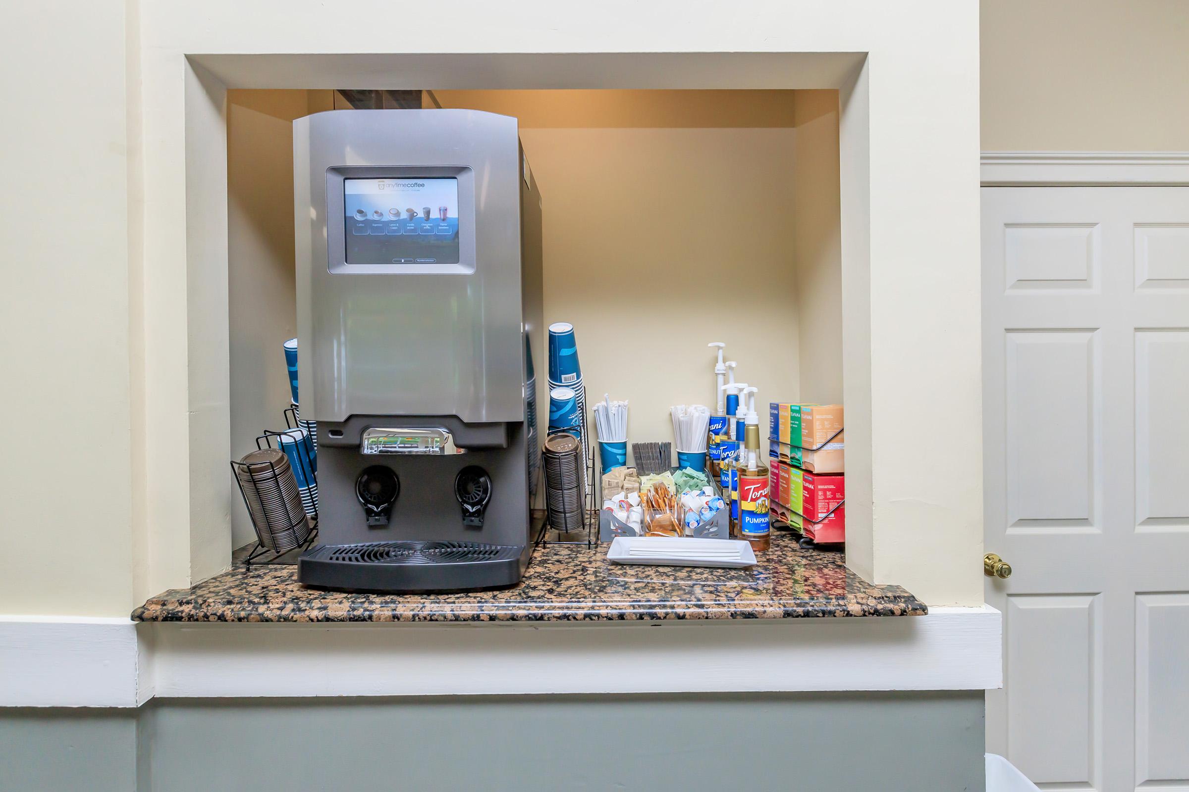 A countertop beverage station featuring a drink dispenser with a digital display, surrounded by cups, napkins, and various condiment containers. The station is set against a light-colored wall, with a white door visible in the background.