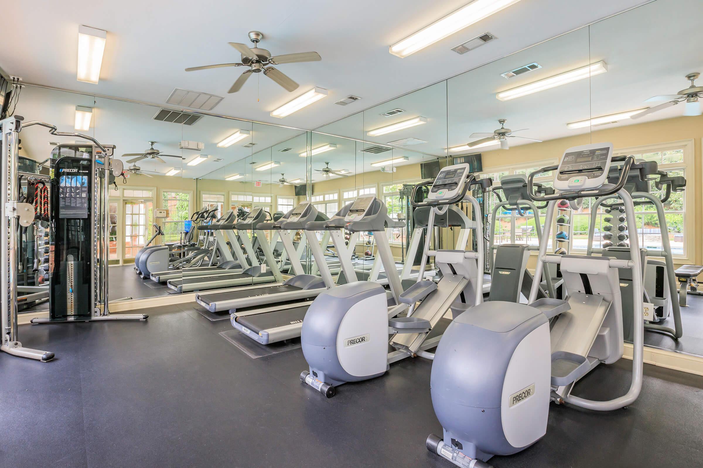 A well-lit fitness center featuring several elliptical machines and treadmills lined up against a mirror wall. The space is equipped with various gym equipment and large windows allowing natural light to fill the room. Ceiling fans are installed for ventilation.