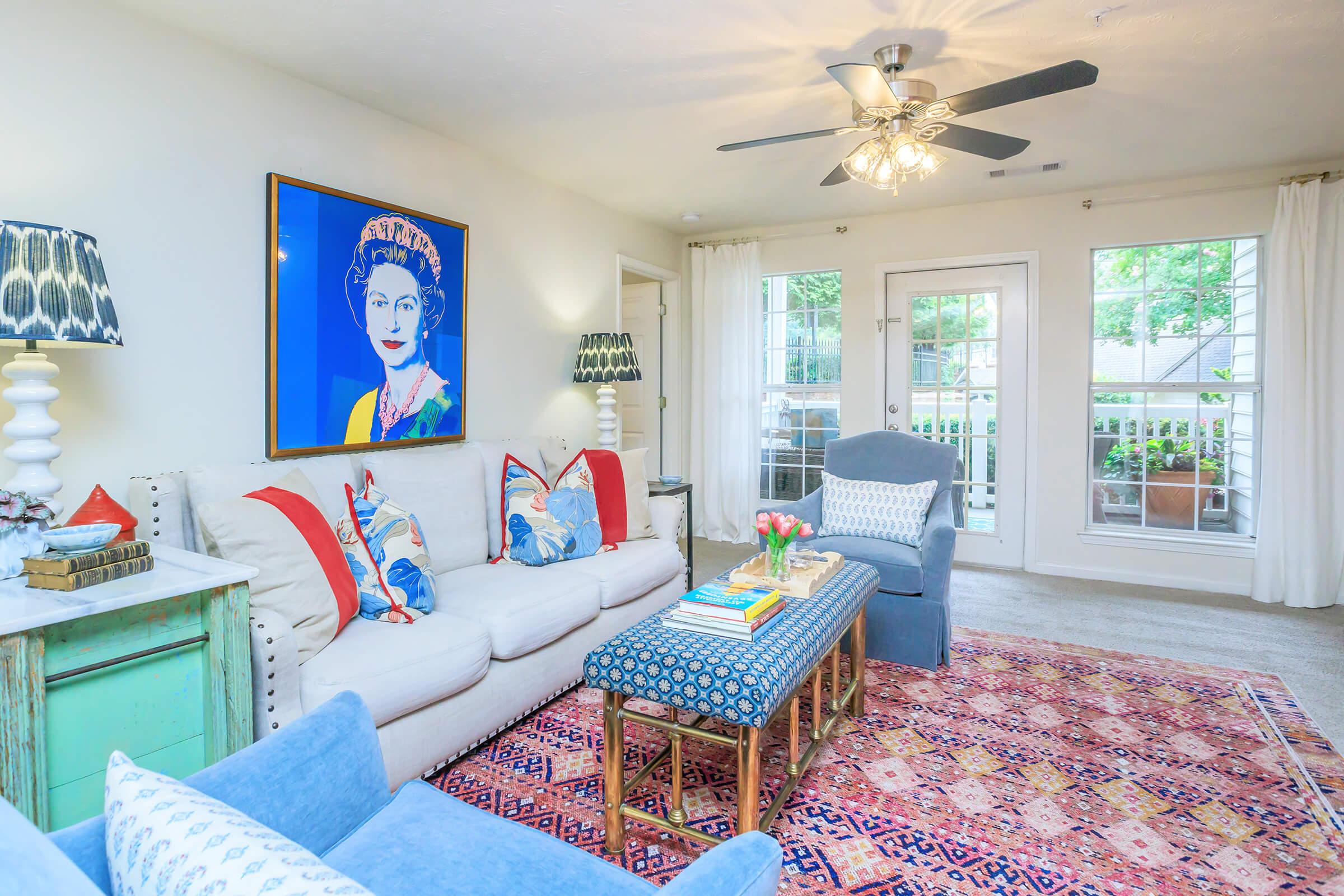 A cozy living room featuring a colorful portrait on the wall, a comfortable white sofa with decorative pillows, a patterned blue coffee table, and a vibrant area rug. Two stylish lamps and large windows bring in natural light, enhancing the warm, inviting atmosphere.