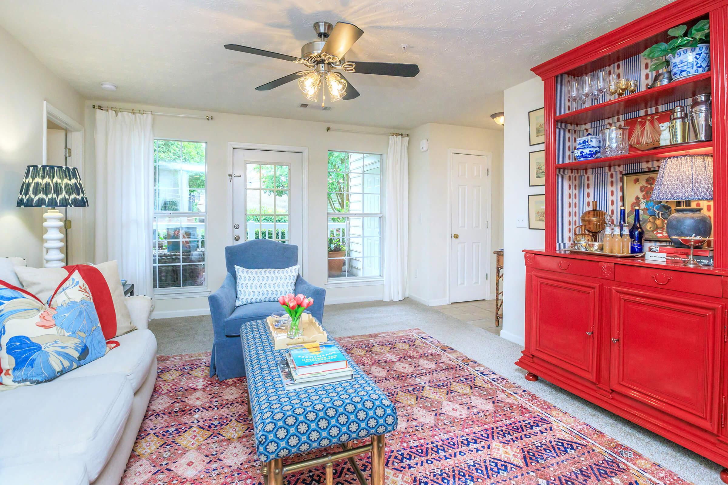 A cozy living room featuring a white sofa adorned with colorful pillows, a blue accent chair, and a patterned area rug. A red wooden cabinet displays decorative items, and large windows allow natural light. A coffee table holds books and a vase with flowers, creating a welcoming atmosphere.