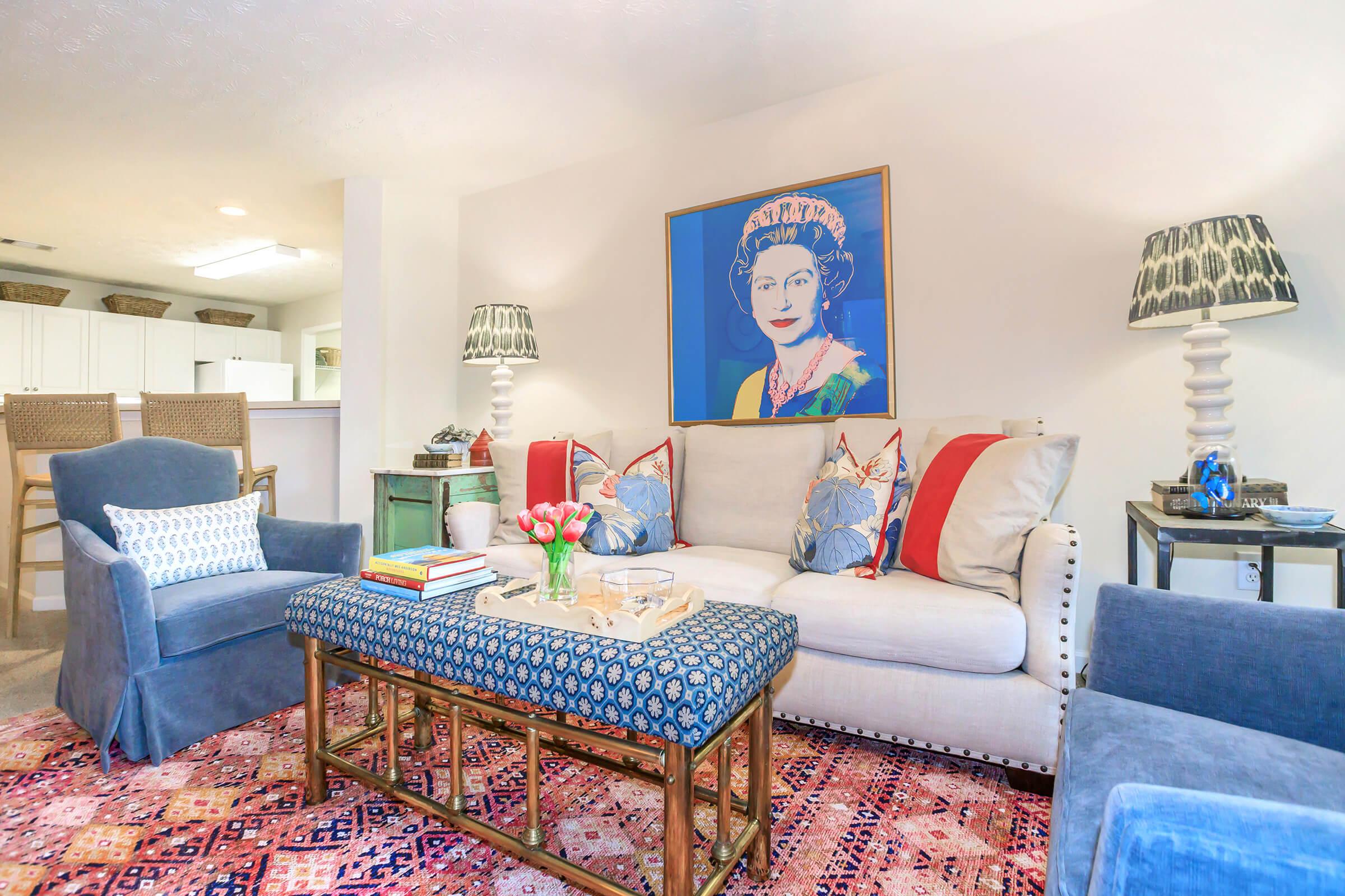 Living room with a light-colored sofa adorned with colorful cushions, a patterned coffee table, and a vibrant area rug. Two chairs flank the sofa, and a large framed portrait of a woman hangs on the wall. Decorative lamps and a vase of flowers complete the cozy, stylish ambiance.