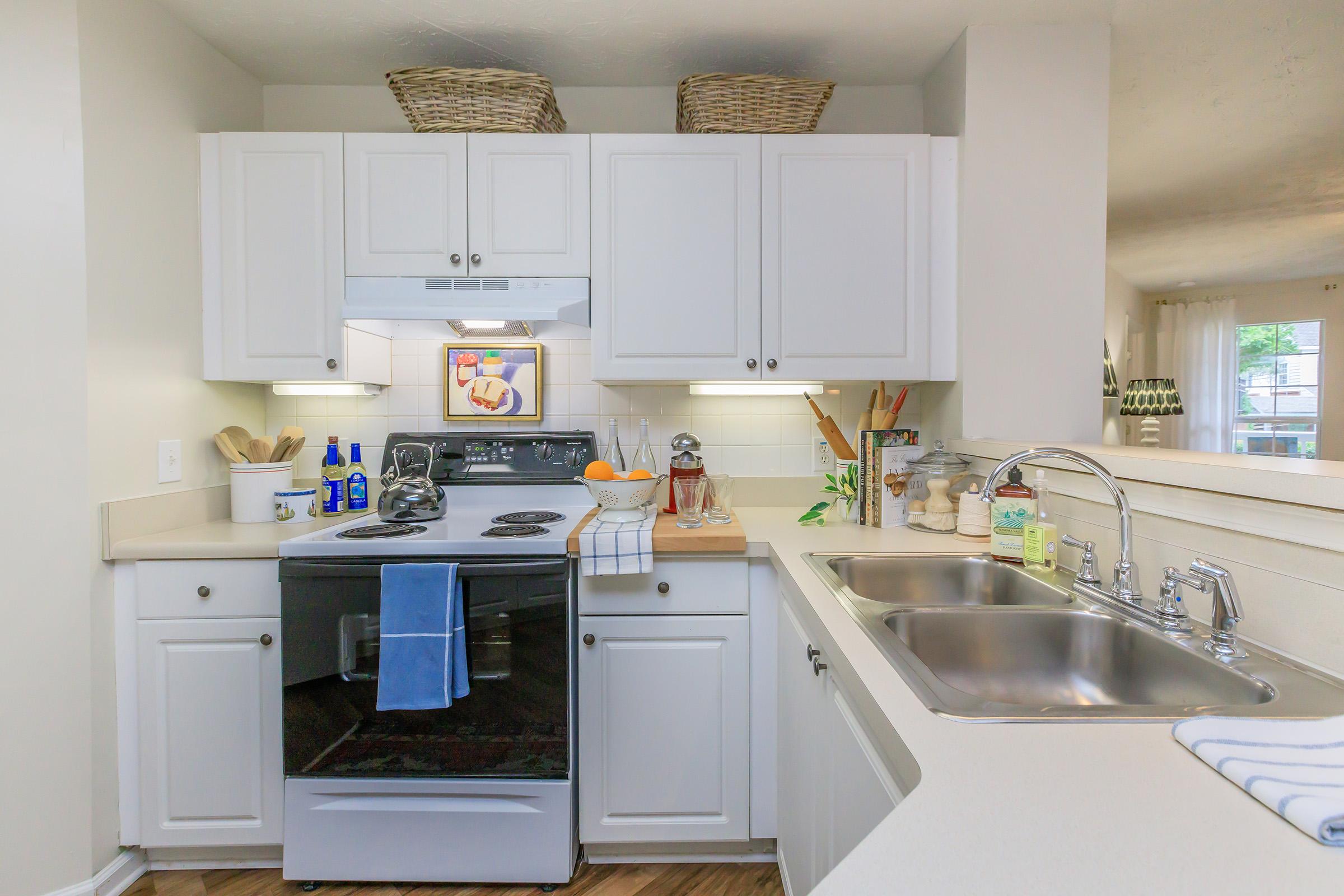 A modern kitchen featuring white cabinetry, a black stove with a blue towel, a double sink with silver faucets, and various kitchen items like bottles and utensils. The countertop is neatly organized, and there are baskets on top of the cabinets for storage. Natural light comes from a nearby window.