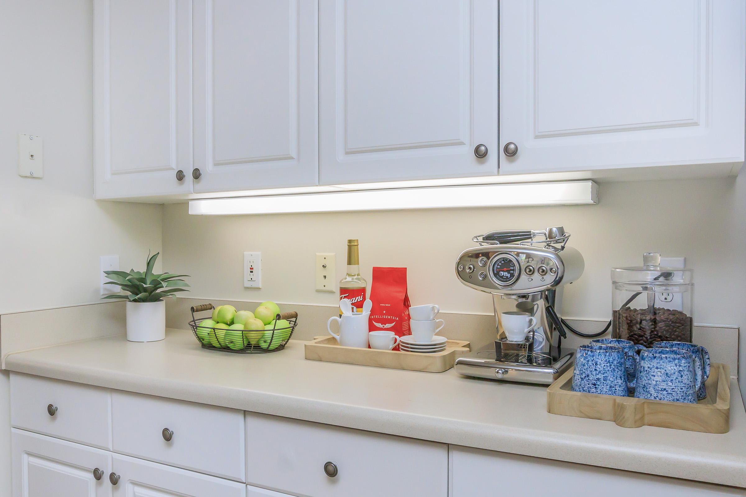 A modern kitchen countertop featuring a coffee machine, a bottle of liquor, a red coffee bag, and two white cups. There's a basket of green apples and a glass container holding coffee beans, all arranged neatly against a backdrop of white cabinets and a lighted shelf.