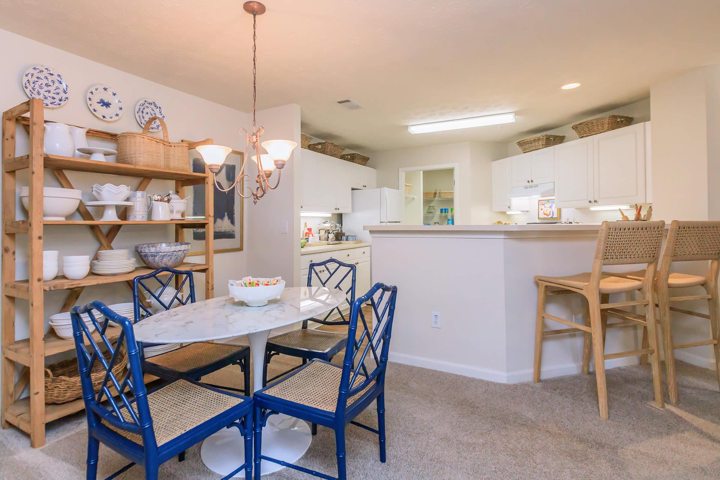 A cozy dining area featuring a round marble table with blue chairs, surrounded by a wooden shelf displaying plates and bowls. In the background, a modern kitchen is visible with white cabinetry and bar seating. Soft lighting from a chandelier adds warmth to the space, which has a neutral carpet.