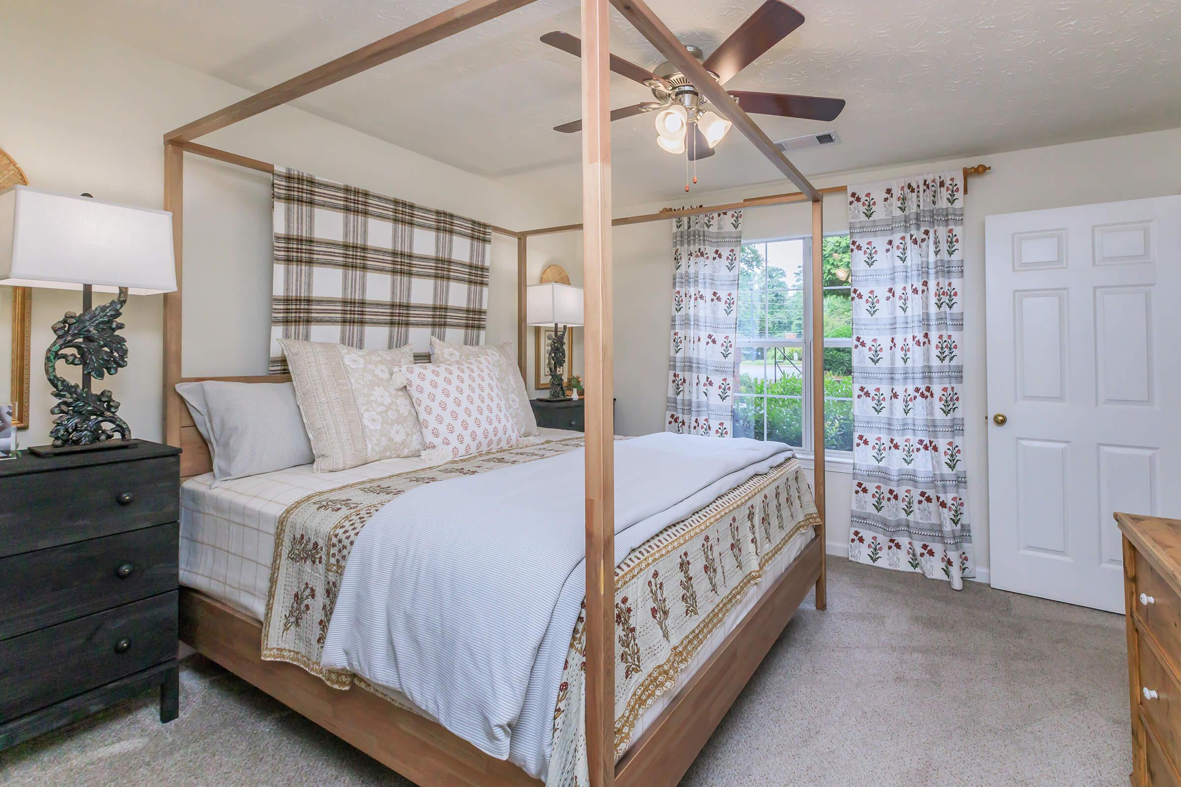A cozy bedroom featuring a four-poster bed with decorative pillows, a patterned throw blanket, and matching bedside lamps. The room has beige walls, a brown dresser, and large windows with patterned curtains, providing natural light and a view of the outdoors.