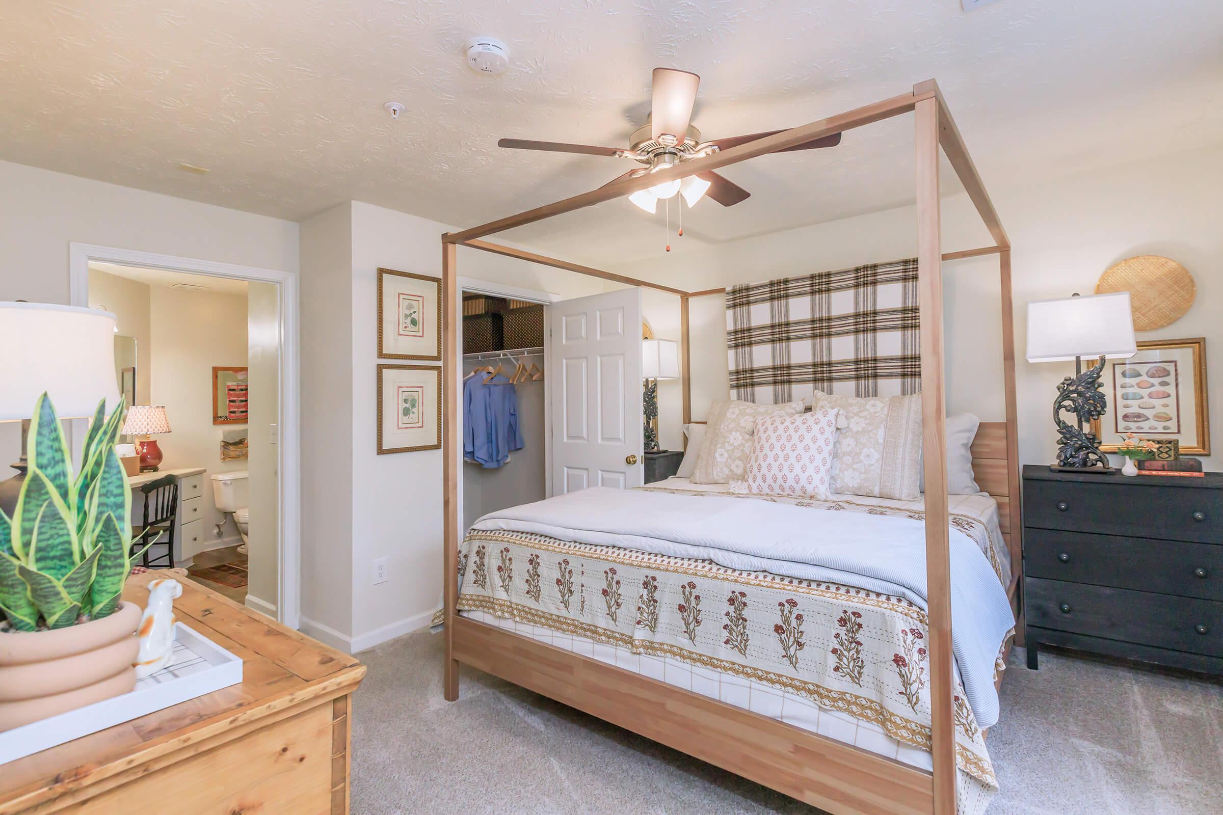 A cozy bedroom featuring a four-poster bed with decorative pillows and a patterned bedspread. The room has beige walls, a ceiling fan, and two bedside lamps. A closet is visible on one side, and a doorway leads to a bathroom. Decorative artwork and a potted plant add charm to the space.
