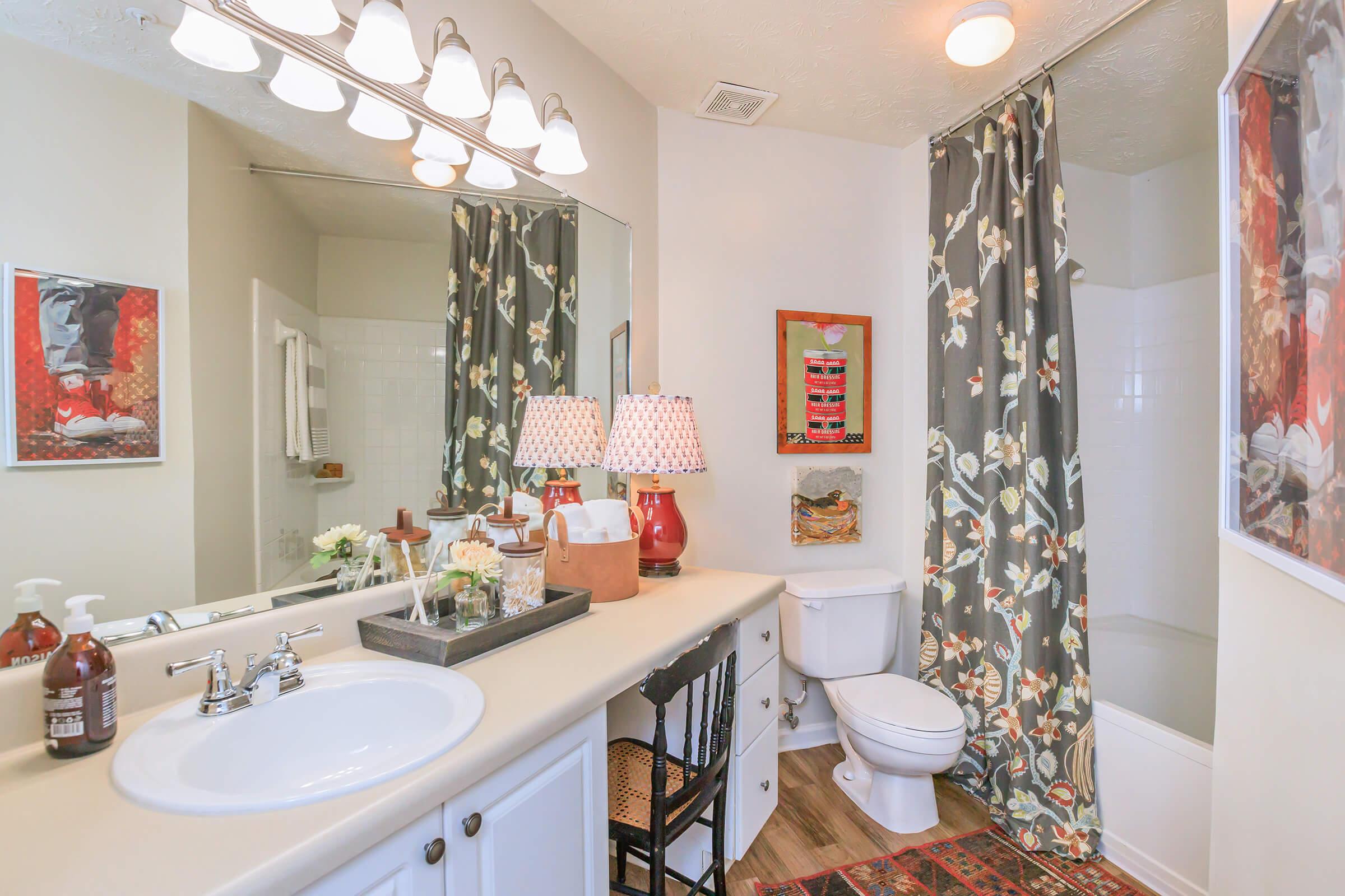 Brightly lit bathroom featuring a double sink vanity with decorative lamps, a colorful shower curtain, and framed artwork on the walls. A towel rack and a small decorative arrangement are visible on the counter, with a toilet and bathtub in view. The overall decor is modern and inviting.