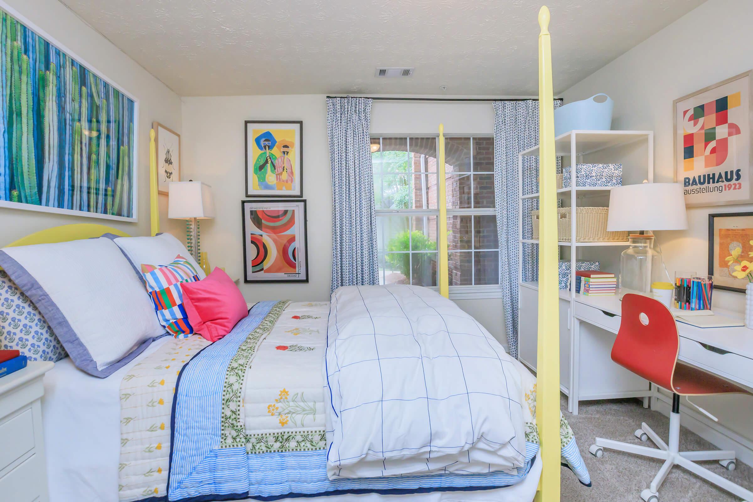A cozy bedroom featuring a neatly made bed with colorful pillows and a quilt, a desk with a red chair, and artwork on the walls. There are large windows with curtains allowing natural light, and a room divider adds privacy. The decor includes vibrant colors and modern design elements.