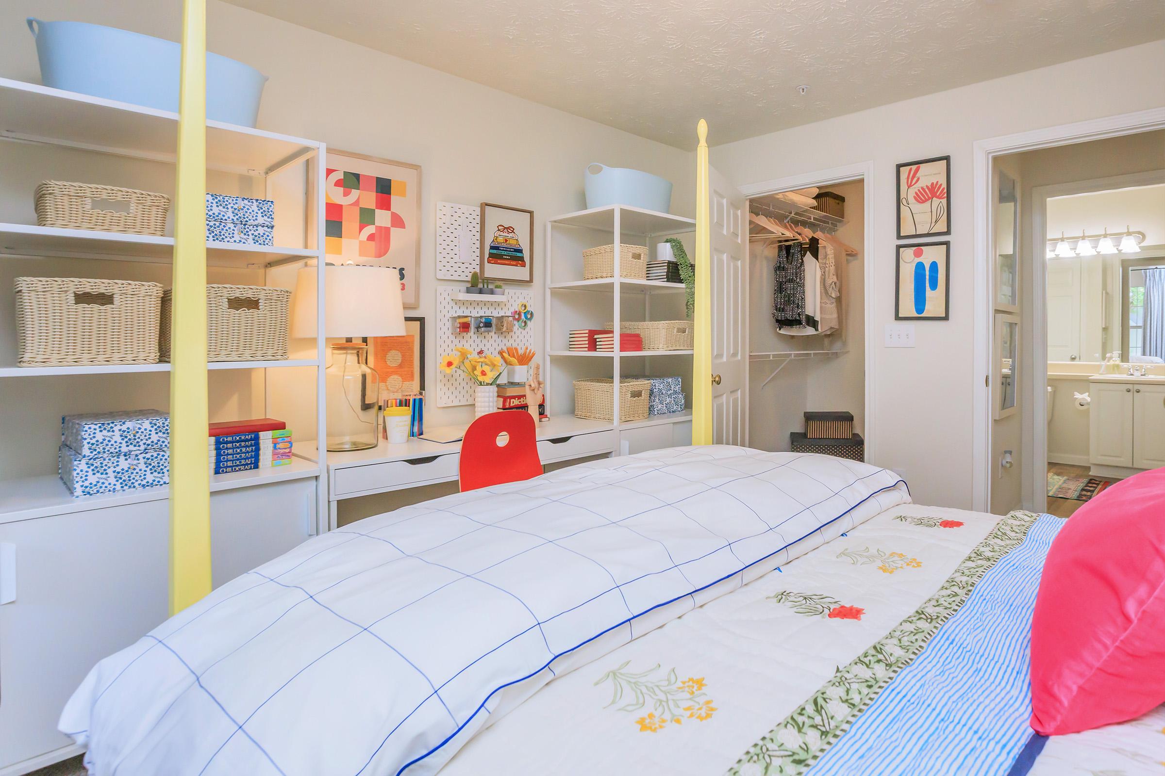 Brightly decorated child’s bedroom featuring a bed with colorful bedding, a desk with a red chair, and open shelves displaying baskets and books. The room is well-lit with a warm ambiance, and a closet door is partially open, revealing additional storage space. Decor includes playful artwork and plants.