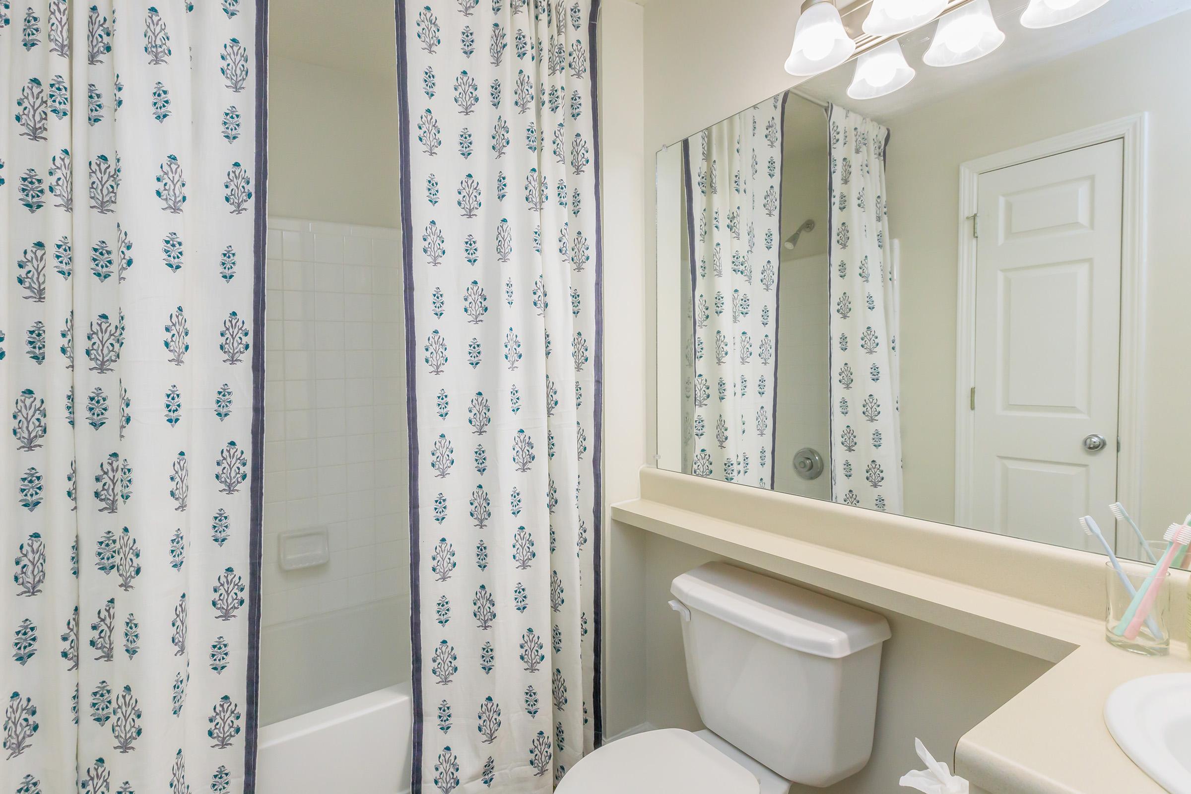 A clean bathroom with a shower area partially covered by a floral shower curtain. There's a countertop with a sink, a mirror reflecting the room, and a bright light fixture above. The walls are painted a light color, and a white door is visible in the background.