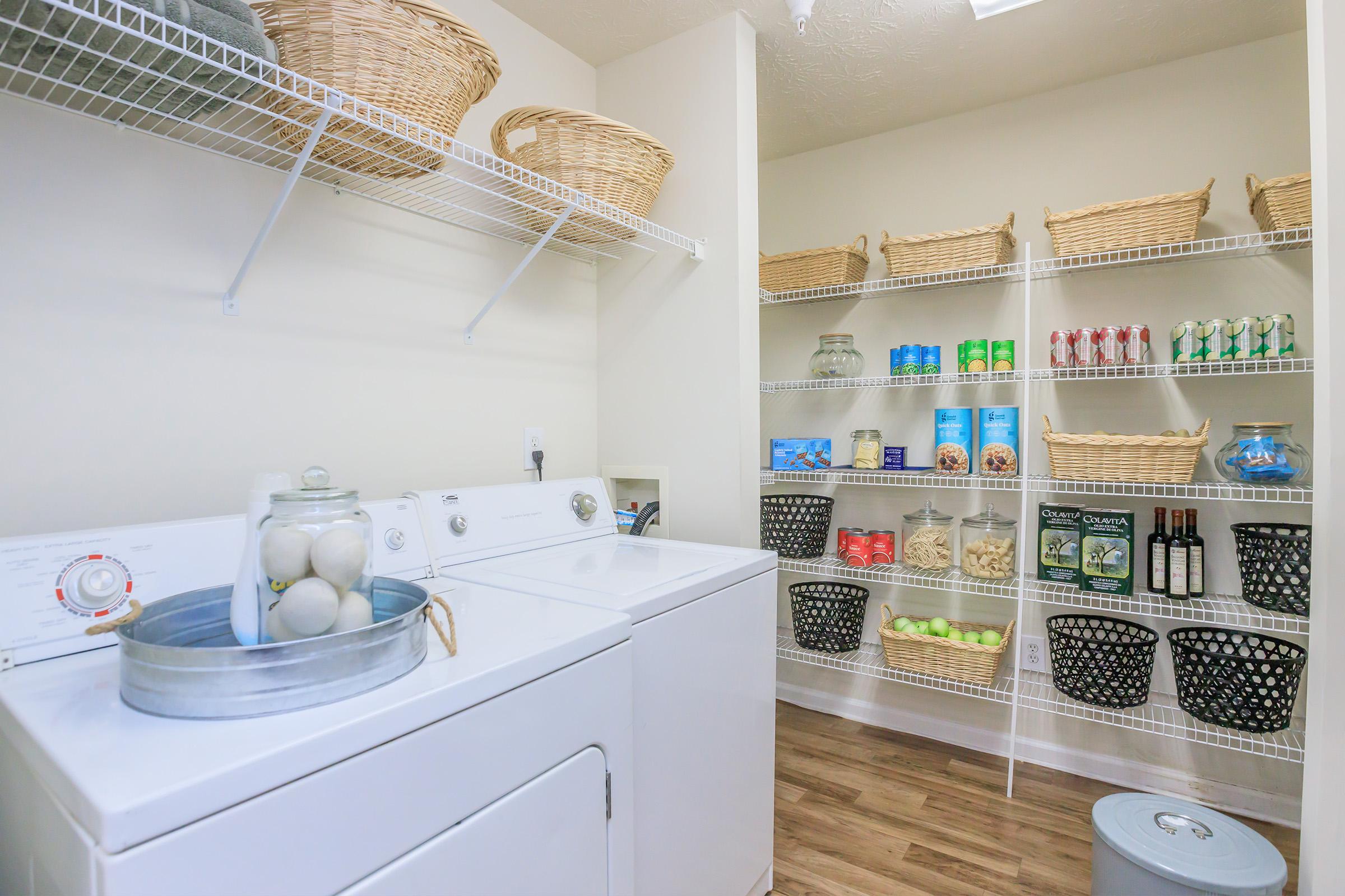 A well-organized laundry room featuring a white washing machine and dryer, with a metallic basket containing laundry items on top. Shelving filled with neatly arranged baskets and containers holds cleaning supplies and other household items. The space is bright and clean with wood-like flooring.